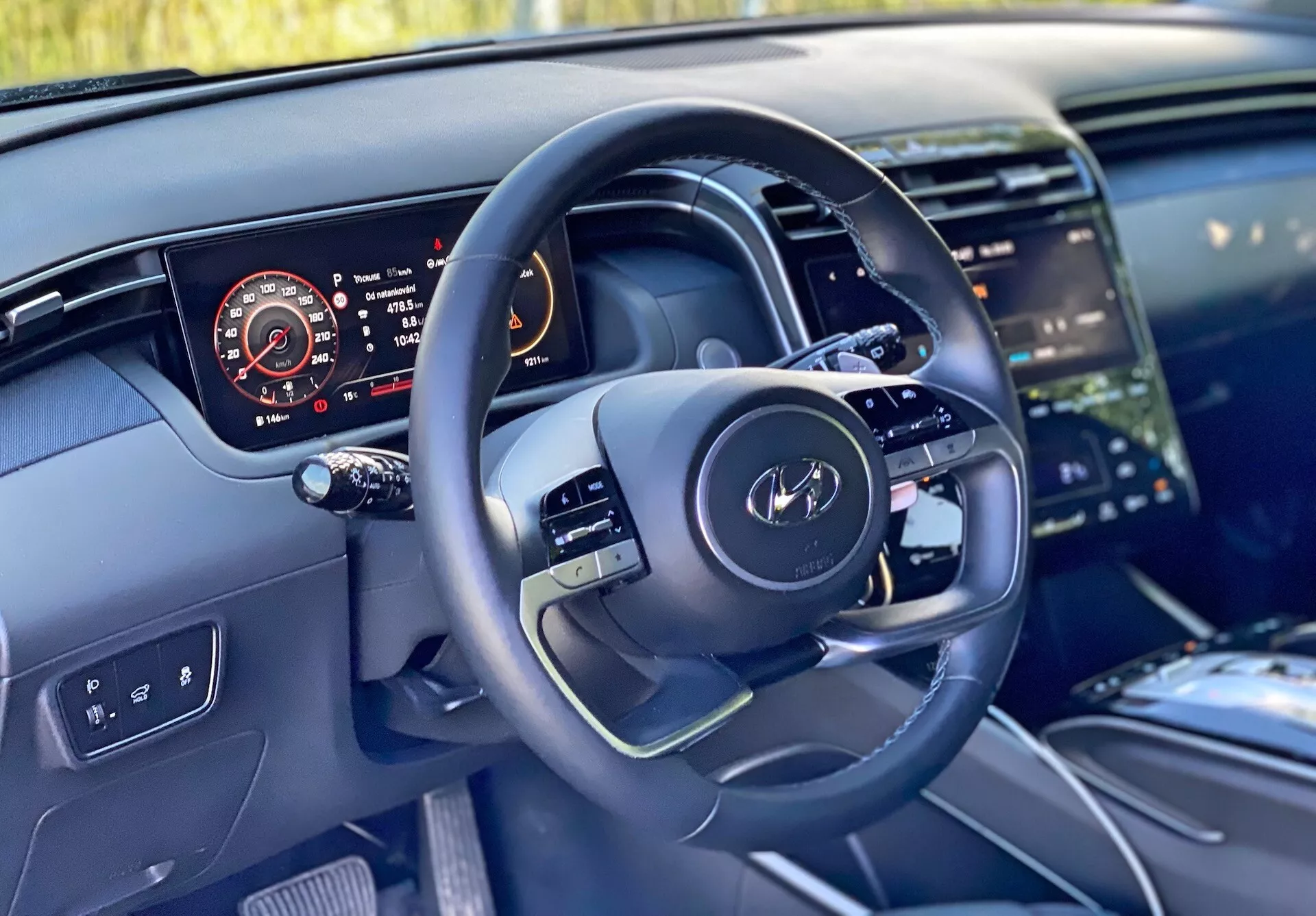 Notranji pogled Hyundai Tucson 2020 iz perspektive voznika, ki prikazuje sodoben volan z večfunkcijskimi kontrolami in digitalni prikazovalnik instrumentov z merilnikom hitrosti, tahometrom, nivojem goriva in temperaturo motorja. Slika poudarja tudi del sredinske konzole z velikim zaslonom na dotik infotainment sistema, čist dizajn armaturne plošče z ambientalno osvetlitvijo ter visoko kakovostnih materialov, ki nakazujejo luksuzne zaključke.