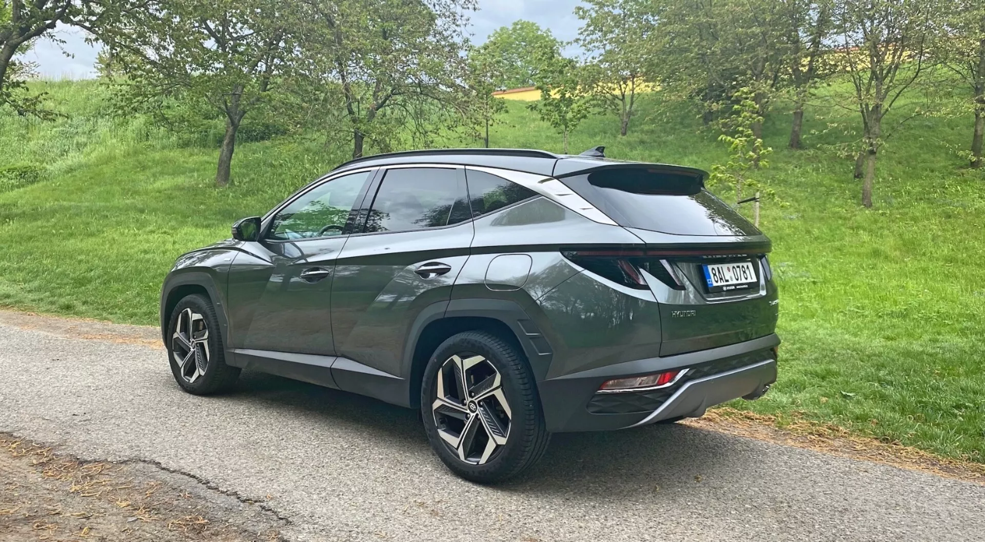 Tričetrtinska zadnja perspektiva Hyundai Tucson 1.6 T-GDi Pure SUV 2020, parkiranega na ozki cesti v podeželskem okolju, prikazuje velike večkrake aluminijaste platišča, temno kovinsko barvo s padajočo strešno linijo, zadnji spojler, zatemnjena stekla na zadnjih oknih, moderne zadnje luči ter dvojni zaključki izpušnih cevi, vgrajeni v zadnji odbijač.