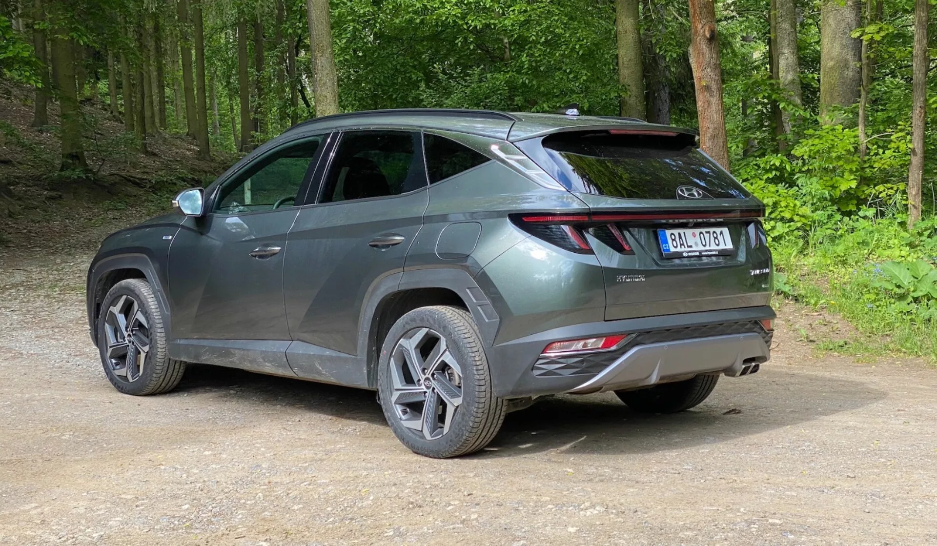 Sodobni SUV parkiran na neasfalitirani poti v gozdnem območju, posnet iz zadnjega tridimenzionalnega kota z rahlega zvišanja. Vozilo ima eleganten, sodoben dizajn z ostrimi linijami, podaljšane zadnje luči, ki zavijajo zadaj na stranice, vidni spojler in odsevnike na zadnjem odbijaču, kar nakazuje napredne varnostne funkcije. Vozilo v kovinsko sivi barvi ima večkrake platišča in cestne pnevmatike, z bleščečo površino, kar kaže, da je novo ali dobro vzdrževano. Okoli avtomobila je zelen, bujen gozd s stisnjeno zemljo in ohlapnim gramozom na tleh.