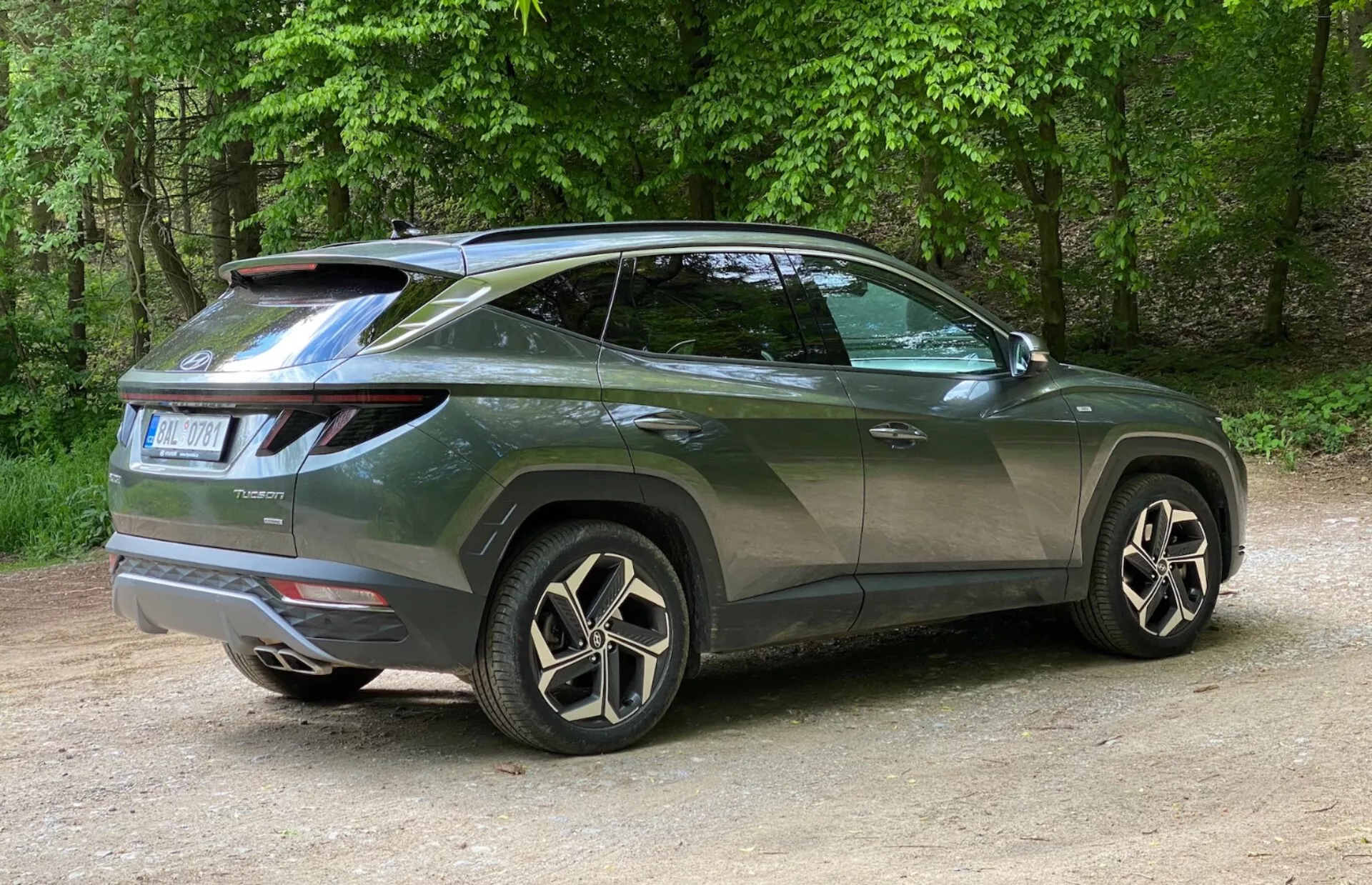 Zadnji pogled treh četrtin modernega SUV-ja Hyundai Tucson 2020 s poudarkom na bočnem profilu in zadnjem dizajnu. Vozilo je postavljeno na levo, fotografija je posneta z desne, s poudarkom na zadnjem desnem kotu. Značilne LED zadnje luči se ovijejo od zadka proti stranem, strešni spojler pa dodaja športni videz. Dvignjena linija pasu oken, poševna strešna linija, zatemnjena okna in kljuke vrat v barvi karoserije poudarjajo videz kupeja. Velika aluminijasta platišča z večkrakim dizajnom in nizkoprofilne pnevmatike povečujejo občutek luksuza. Manjše podrobnosti, kot so zadnji brisalec in obrobe blatnikov, pomenijo praktičnost in robustnost. Avto je postavljen na gramozu v naravnem okolju z drevesi v ozadju.