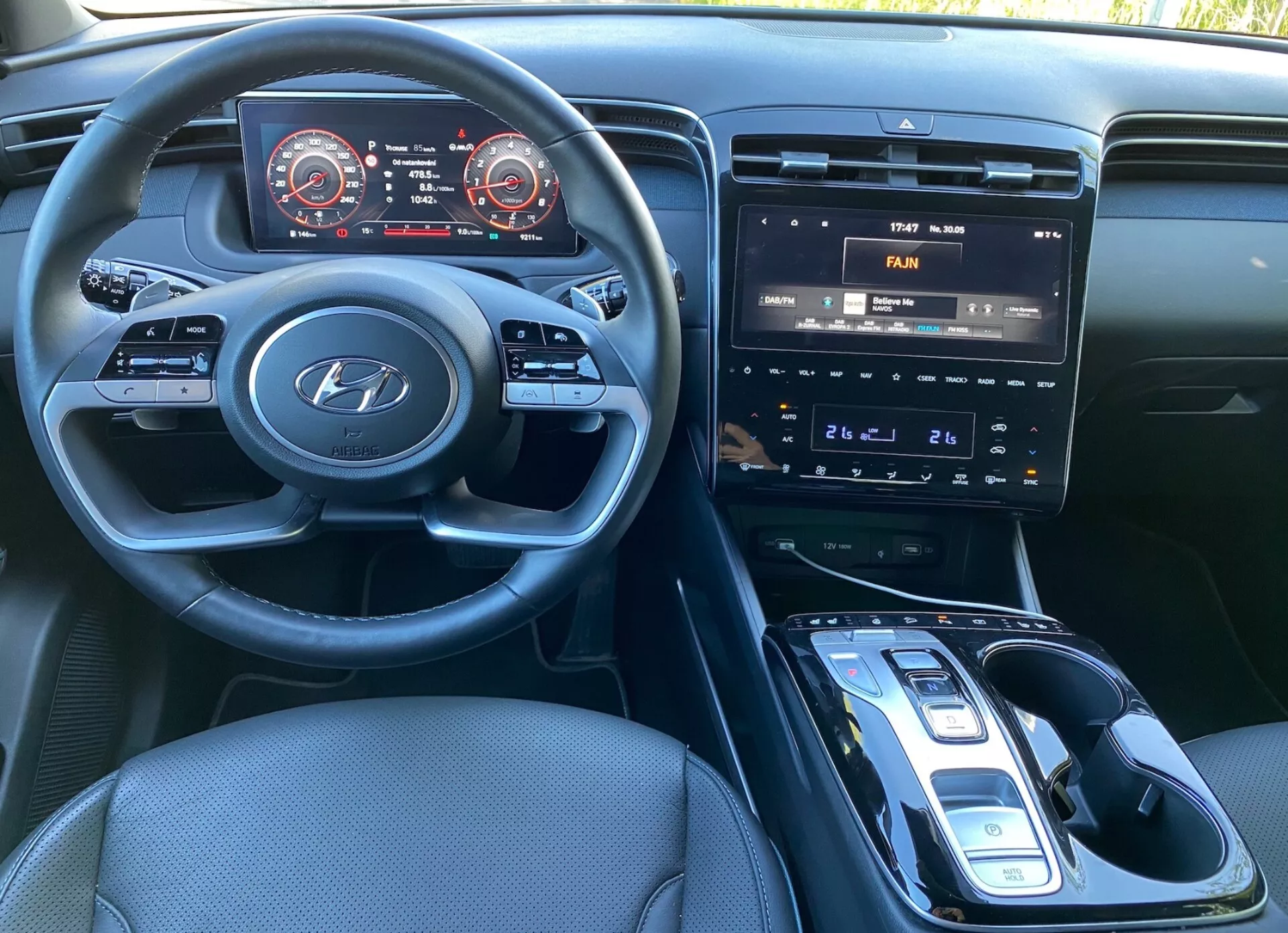 Notranji pogled Hyundai Tucson 2020 iz voznikovega sedeža, ki prikazuje volan z logotipom Hyundai in nameščenimi krmilniki za audio, telefon in nastavitve tempomata. Armaturna plošča vključuje digitalno instrumentno ploščo s hitrostjo in merilnikom vrtljajev z rdečimi poudarki. Sredinska konzola ima informacijski sistem z zaslonom na dotik, ki prikazuje krmiljenje klimatske naprave, fizične gumbe in gumbe za nadzor temperature ter avtomatski menjalnik z elektronskim izbirnikom. Vidna je potencialna brezžična polnilna pločica, temna tkaninska oblazinjenja in dobro osvetljena kabina.
