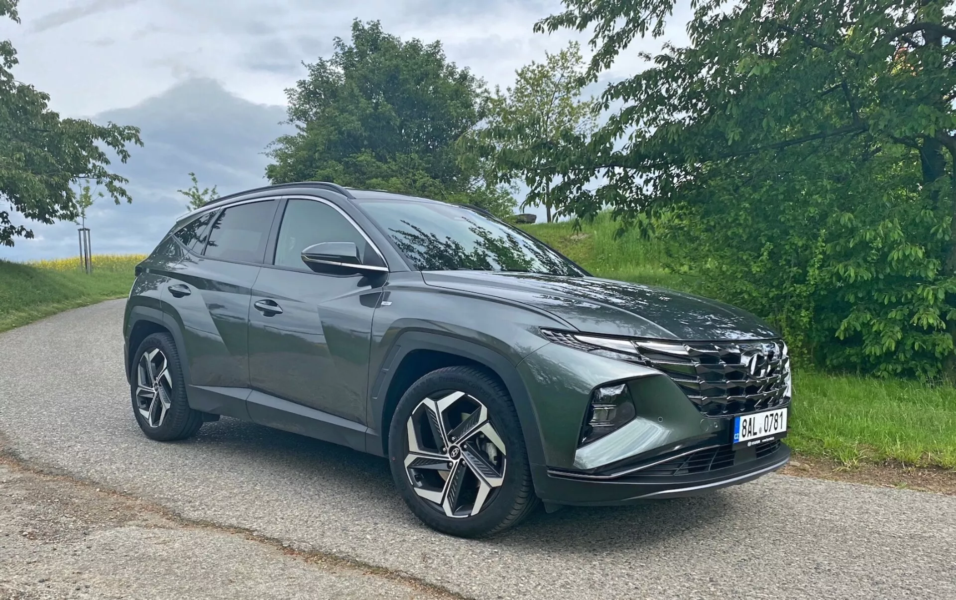 Slika prikazuje sodoben SUV Hyundai Tucson 1.6 T-GDi Pure, parkiran na asfaltirani cesti s naravno pokrajino v ozadju. Avto je fotografiran iz sprednjega kota treh četrtin, zaradi česar so vidni izrazita rešetka, elegantni žarometi, sprednji odbijač z meglenkami ali zračnimi režami, velika aluminijasta kolesa z večkrakim dizajnom in stranska ogledala. Silhueta SUV-ja se proti zadnjemu delu spušča, kar kaže na športno, aerodinamično zasnovo. Posneta v mehkih dnevnih svetlobnih pogojih, fotografija poudarja barvo in oblikovne podrobnosti vozila brez ostrih senc.