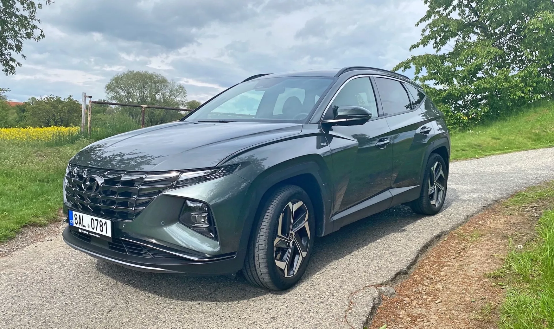 Hyundai Tucson 2020 1.6 T-GDI Pure SUV, prikazan iz prednjega pol stranskega kota na tlakovanem cestišču s travo in drevesi v ozadju. Slika prikazuje rešetko vozila, ostre LED žaromete, platišča iz lahke litine, stranska ogledala z vgrajenimi smerniki ter temno kovinsko barvo. Vidna so voznikova prednja vrata in zadnja vrata. Fotografija je bila posneta podnevi v oblačnih svetlobnih pogojih.