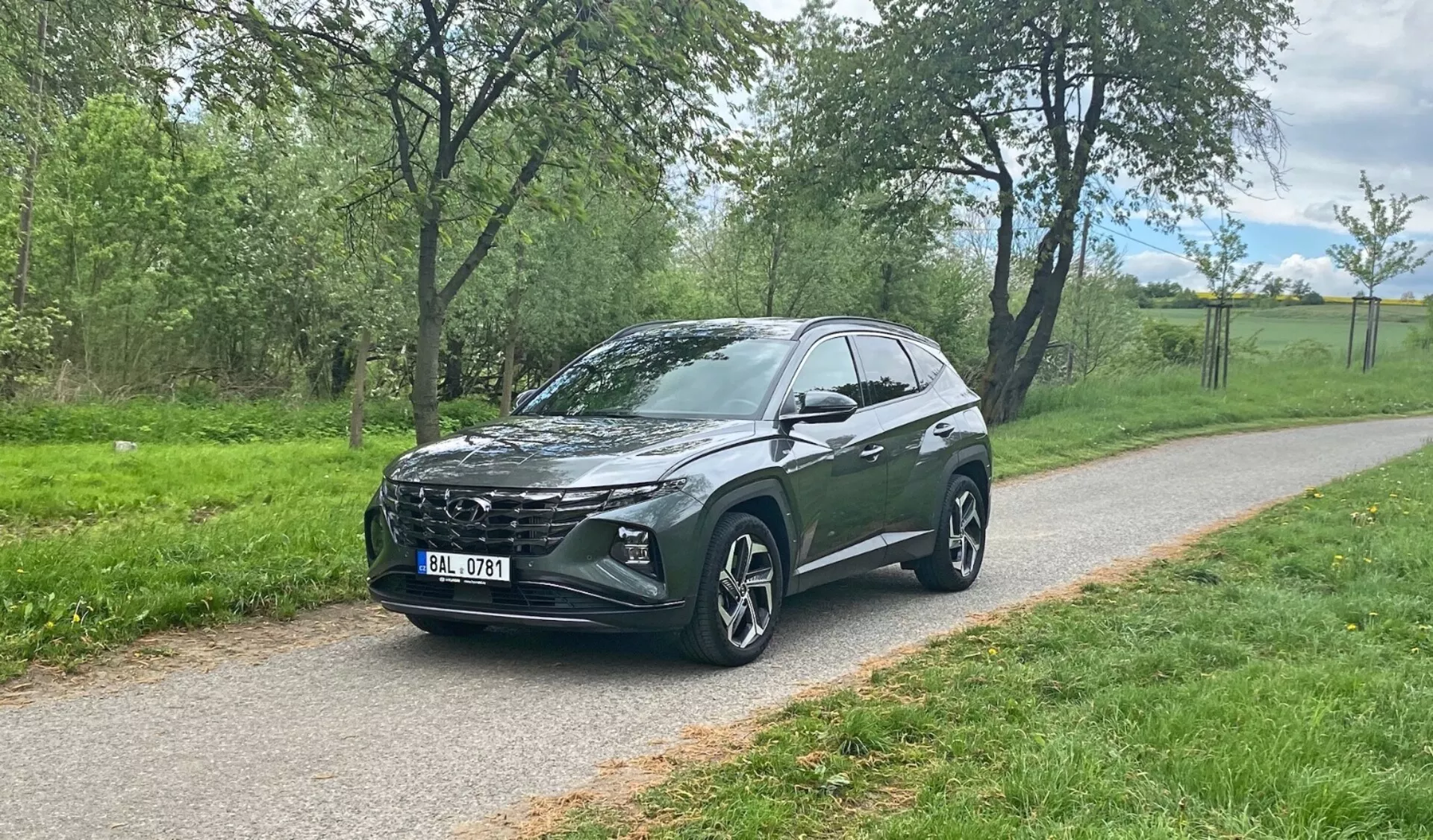 Tričetrtinski sprednji pogled na crossover SUV Hyundai Tucson 2020, parkiran na tlakovanem poti z zelenjem v ozadju, prikazujejoč značilno sprednjo masko vozila, elegantne LED žaromete, velika večkraka aluminijasta kolesa, nagnjeno strešno linijo, visoko višino od tal, integrirana stranska ogledala, zatemnjena stekla in stilizirane oblike karoserije.