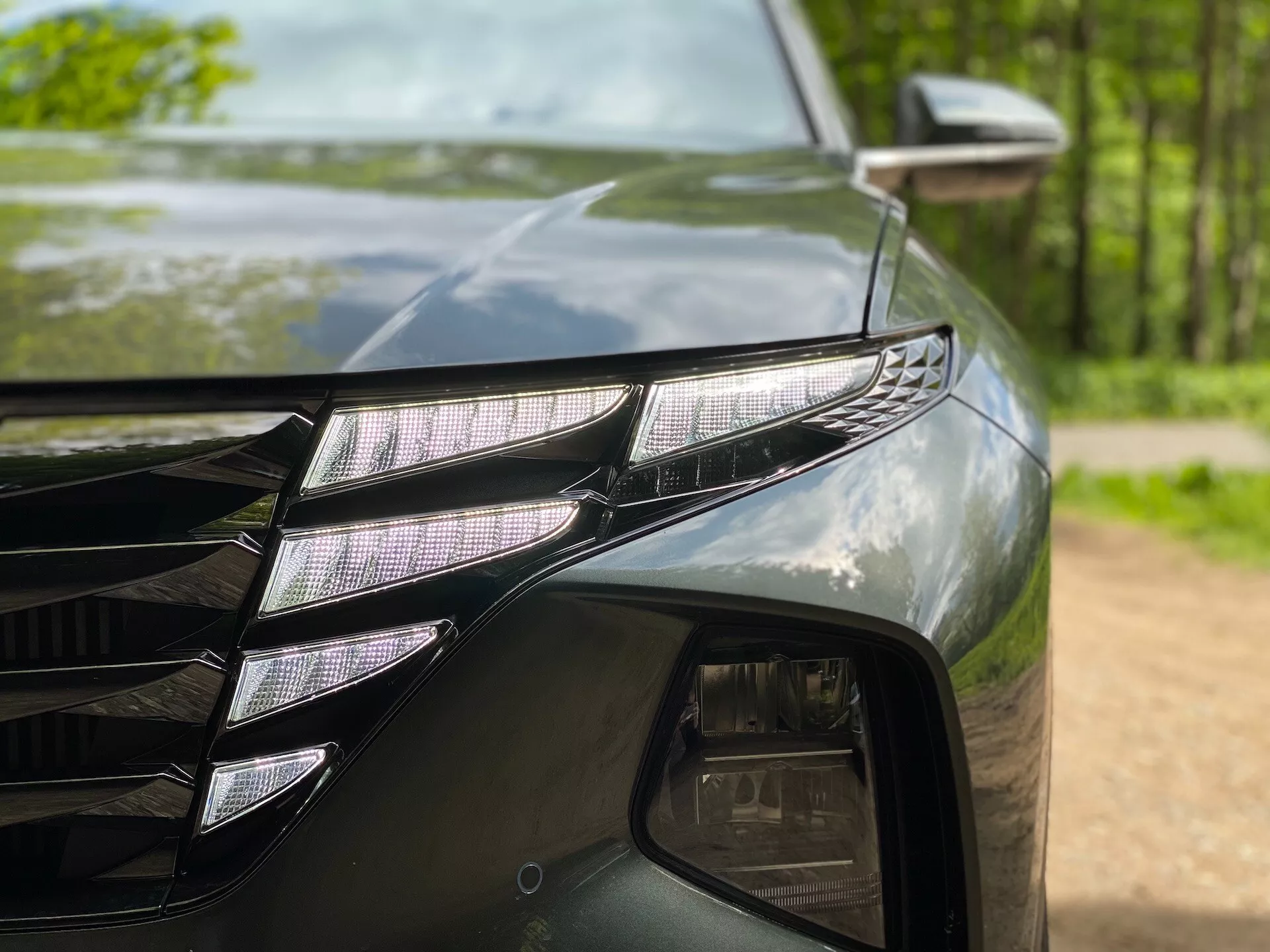 Podroben posnetek leve prednje strani Hyundai Tucson 2020 (Hyundai-Tucson-1-6-T-GDi-Pure), ki prikazuje LED žaromete z individualnimi LED projektorji in značilnim LED DRL trakom. Temna, sijoča barva odseva okoliška drevesa. Viden je tudi del mreže z ostrimi linijami in stransko ogledalo s smernikov indikatorjem.