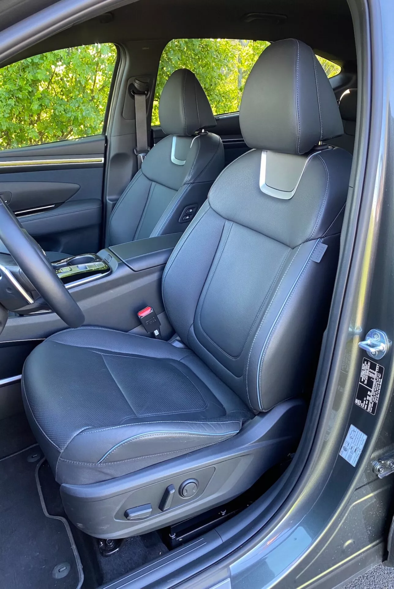 Notranjost Hyundai Tucson 2020 1.6 T-GDI Pure, ki prikazuje sprednji sedež potnika s črnim usnjenim oblazinjenjem, podrobnimi šivi in ​​krmilniki za električno nastavitev. Vidna sredinska konzola s držalom za pijačo, vratna plošča z ujemajočimi se materiali, ročaj, predal in mrežica zvočnika, ki nakazujeta napreden avdio sistem. Delno vidna armaturna plošča in predal za rokavice. Preproga kot talna preproga in rahle sončne odseve na vratni plošči.