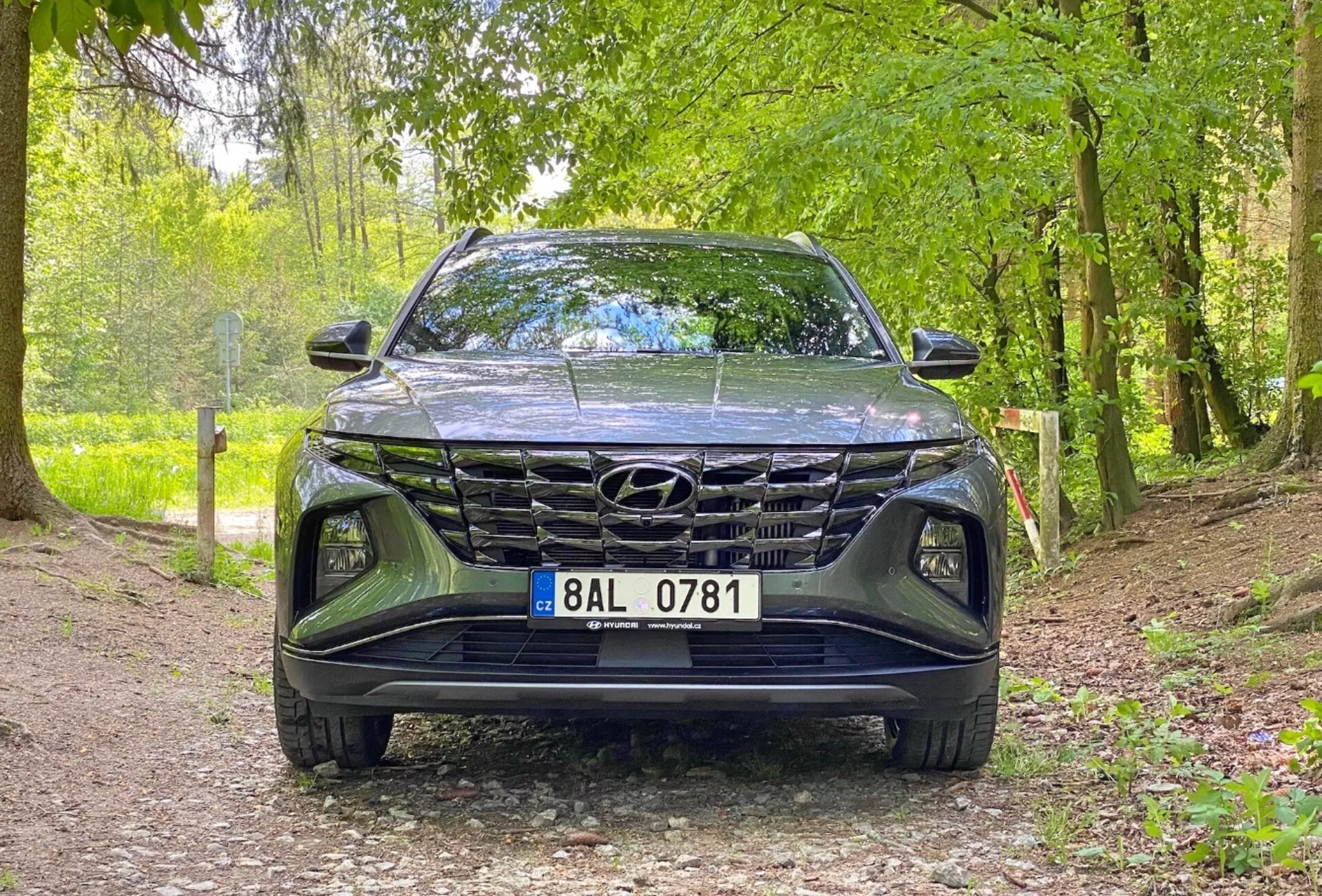 Pogled z sprednje strani na vozilo Hyundai Tucson 1.6 T-GDi Pure 2020, ki prikazuje nizkokotni posnetek, osredotočen na mrežico, sprednji odbijač in območje pokrova motorja. Avto je parkiran na gramozni poti v naravnem okolju, kar nakazuje na primernost za zunanje okolje. Ključne lastnosti vključujejo izrazito obliko mrežice, elegantne LED žaromete in vidno značko Hyundai.