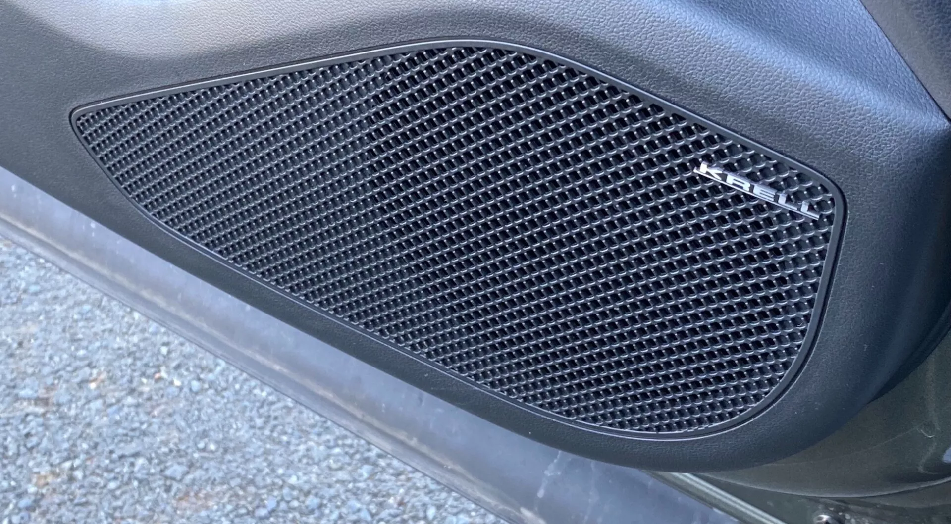 V bližnjem stranskem pogledu Hyundai Tucson 2020 1.6 T-GDI Pure, se osredotoča na stransko rešetko z mrežastim vzorcem v obliki diamantov. Rešetka, vgrajena v oblikovan obris, je oblikovana v karoserijski panel avtomobila z mat končno obdelavo, obkrožena z gladko, ukrivljeno površino. Slika je posneta pod poševnim kotom od spodaj navzgor, prikazuje naravno zunanjo osvetlitev s koščki asfalta in cestnimi smetmi, kar izboljšuje eleganten, zmogljiv videz.