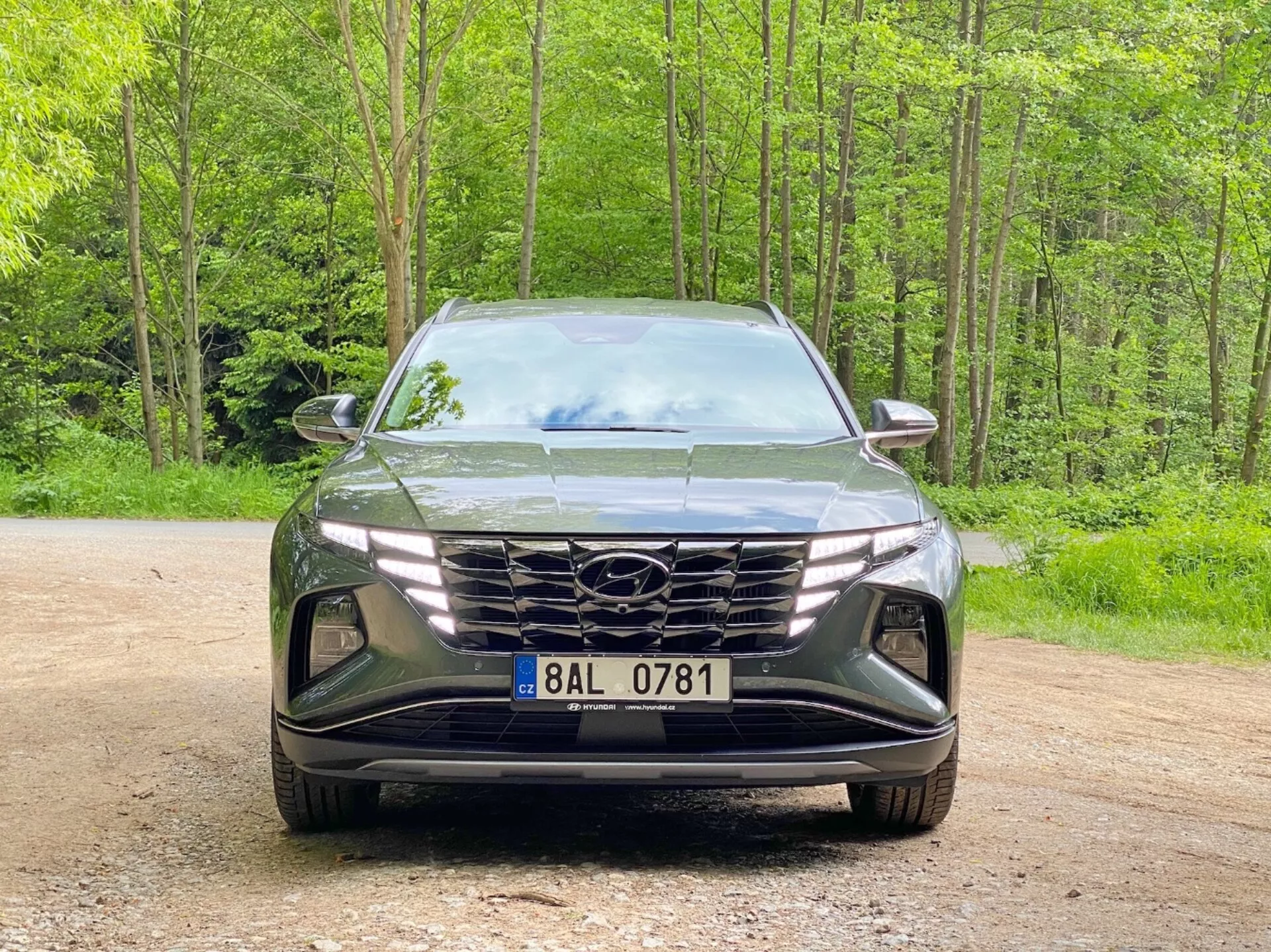 Sprednji pogled na Hyundai Tucson 2020 (Hyundai Tucson 1.6 T-GDI Pure) moderno SUV parkirano na gramozni poti z gozdnim ozadjem. Slika zajame elegantno, agresivno oblikovanje z veliko geometrijsko mrežo, ki vsebuje logotip proizvajalca, ozke kotne LED žaromete in izklesan sprednji odbijač z odprtinami za zrak in režami za meglenke, kar poudarja napredno aerodinamiko in svetlobno tehnologijo.