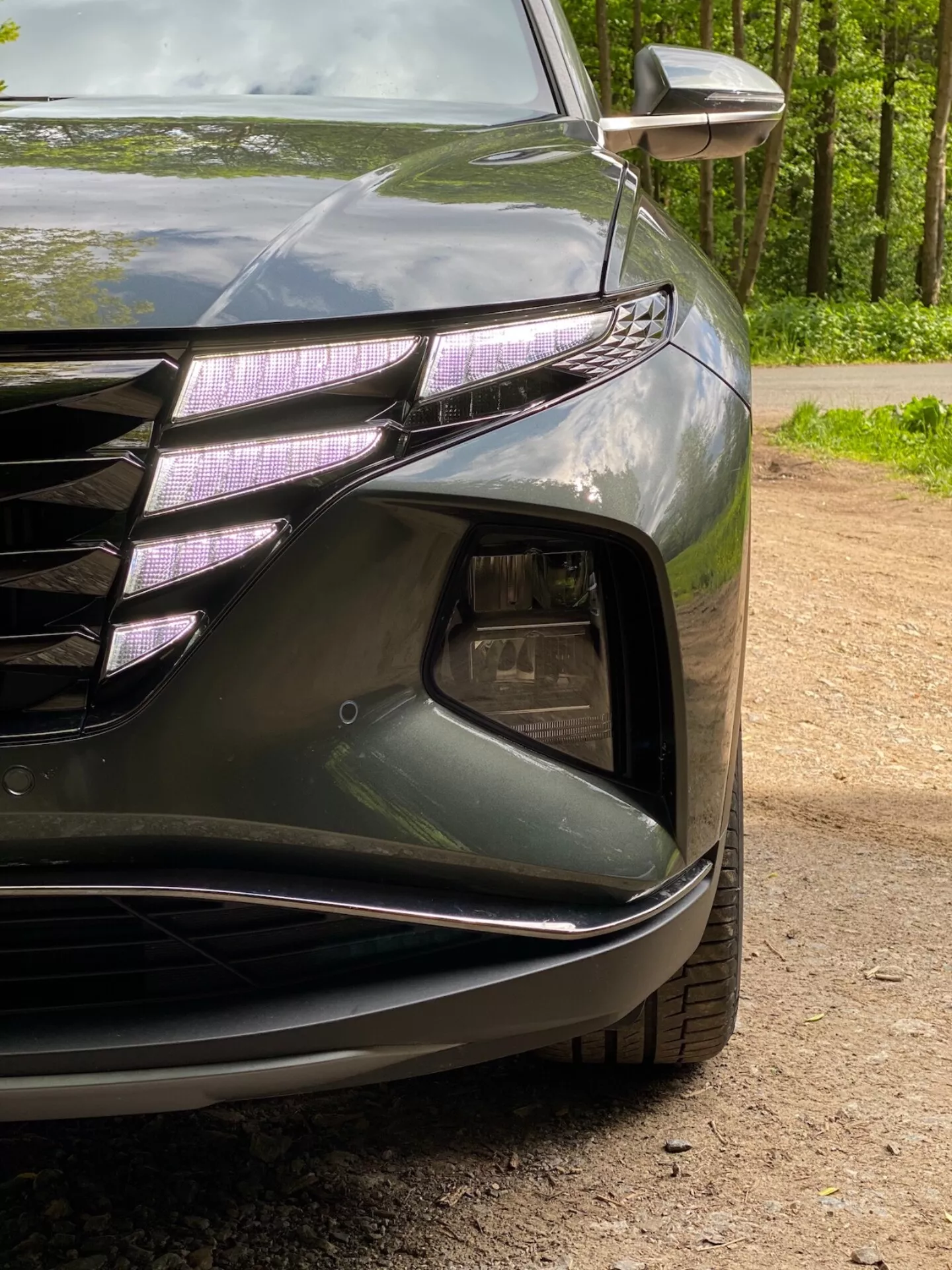 Primeren pogled na prednji levi kot Hyundai Tucson 2020 1.6 T-GDI Pure, ki prikazuje žaromet z LED dnevnimi lučmi in projektorsko lečo, del sprednje rešetke s horizontalnimi linijami in kromiranimi poudarki, sekcijo za dovod zraka z ohišjem za meglenke, skulpturiran sprednji odbijač z gladkimi linijami in vgrajenim smernikom v zunanjih ogledalih. Temna, sijoča barva poudarja aerodinamične krivulje avtomobila.