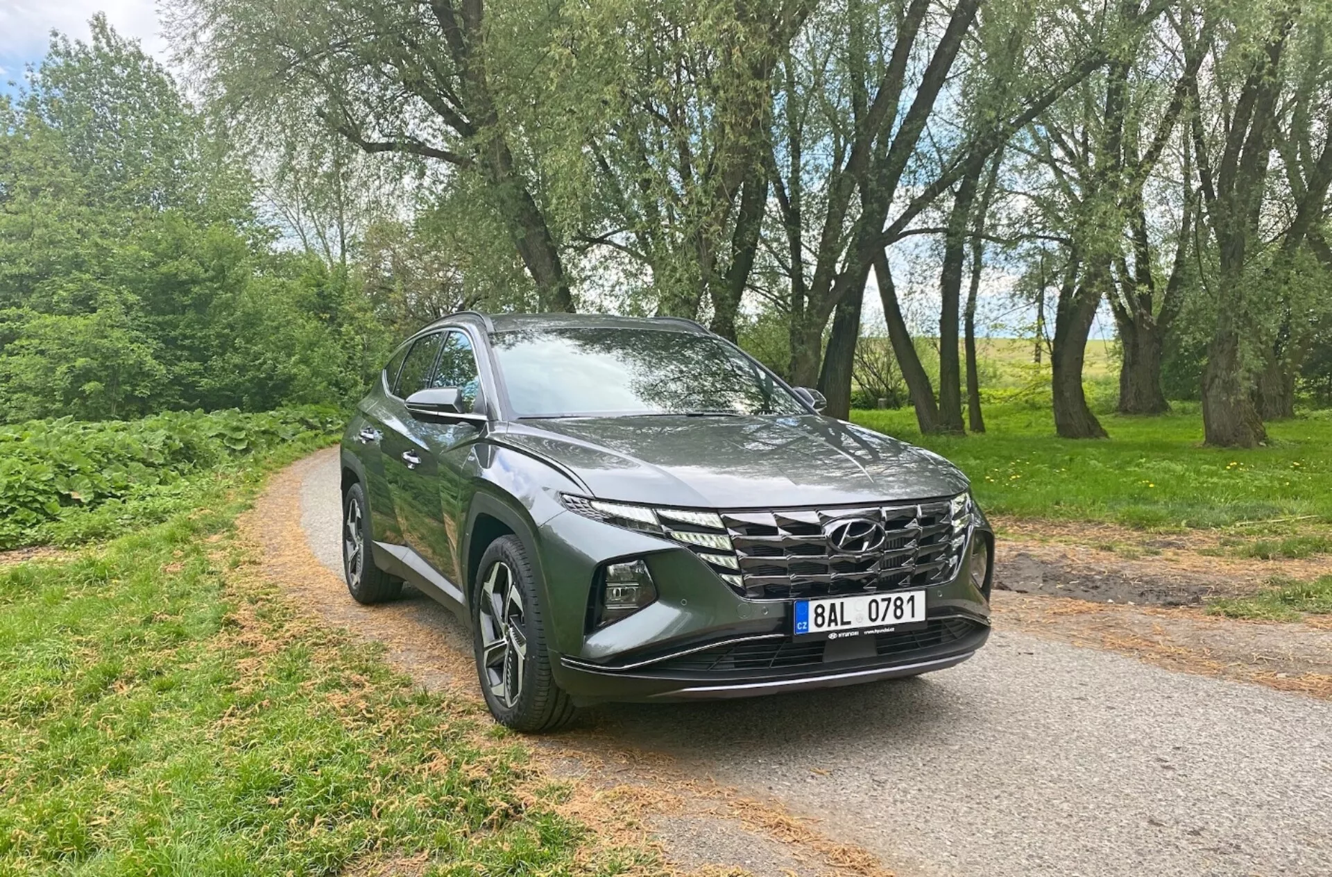 Sprednji tričetrtinski pogled na Hyundai Tucson 1.6 T-GDi Pure 2020, ki prikazuje sodobne SUV lastnosti, kot so elegantni LED žarometi, izrazita geometrijska sprednja rešetka, aerodinamičen pokrov motorja, aluminijasta platišča, stranska ogledala z vgrajenimi smerniki in kovinska barva, v ozadju ozke asfaltirane ceste z drevesi, kar poudarja njegovo vsestranskost in privlačnost tako za mestno kot podeželsko okolje.
