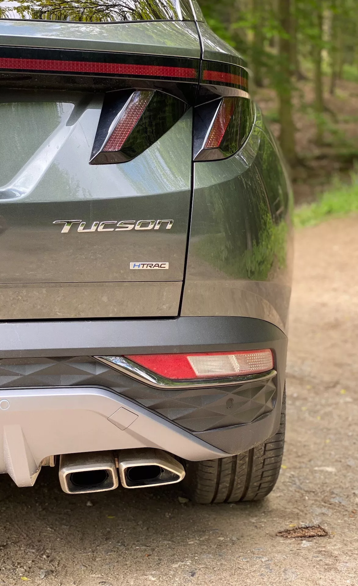 Delni zadnji pogled modela Hyundai Tucson 2020, ki kaže, da gre za različico 1.6 T-GDi Pure, ki prikazuje desno stran odbijača, del zadnje luči, zadnji spojler z LED zavorno lučjo, dvojne izpušne cevi in oznako modela z imenom 'Tucson' in motorno tehnologijo 'T-GDi'.