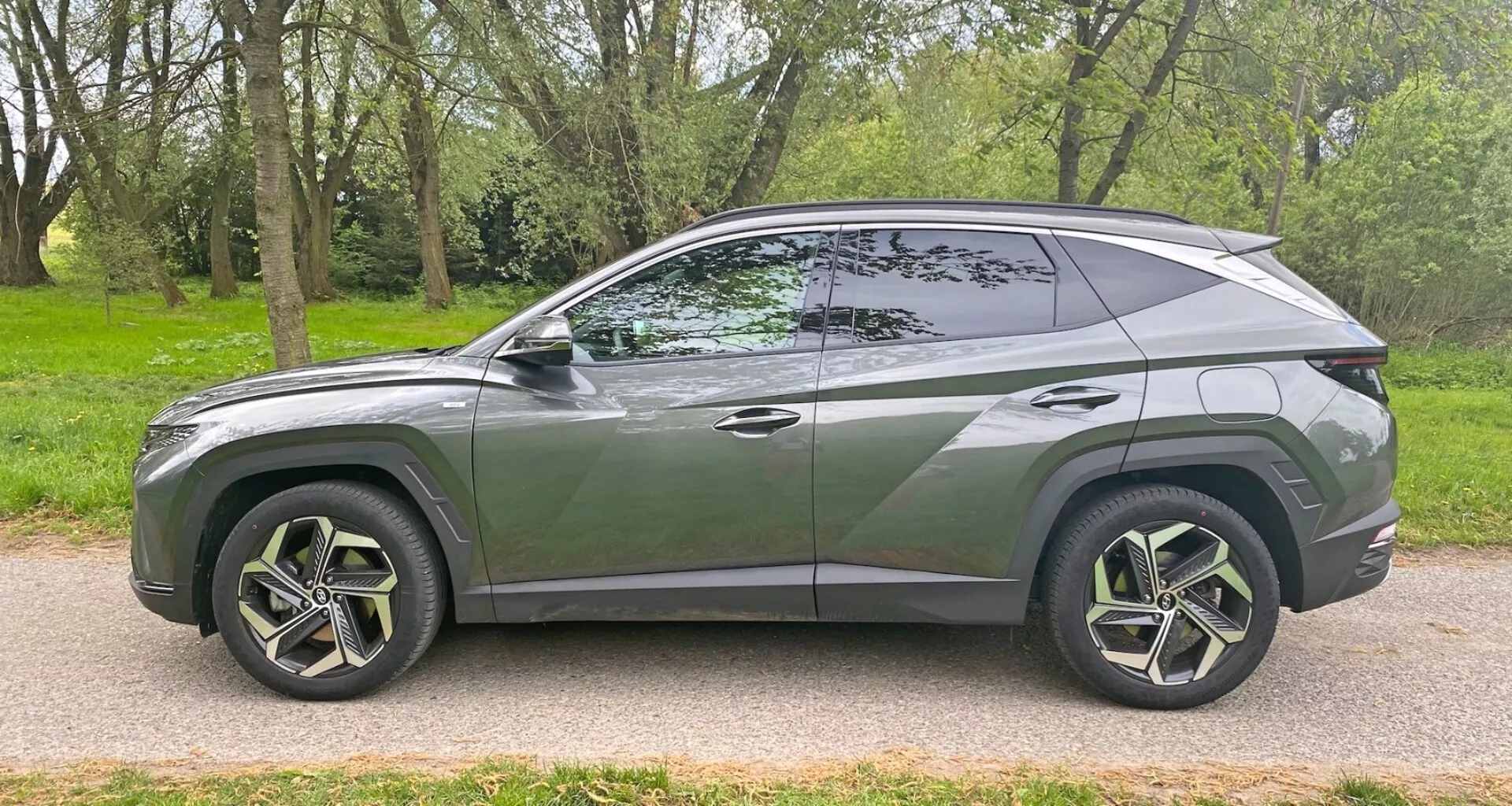 Stranski profil sodobnega SUV Hyundai Tucson 1.6 T-GDI Pure 2020 parkiranega na tlakovanih cestah poleg travnate površine z drevesi. Pokaže eleganten, aerodinamičen dizajn s padajočo strešno linijo, izrazito karoserijsko linijo, velikimi multi-kraki iz lahke zlitine s pnevmatikami z nizkim profilom, zatemnjenimi okni z vidnim B-stebrom, sploščenimi ročaji vrat, stranskimi ogledali barve karoserije in kromiranim obrobkom vzdolž okenskega roba. Vozilo zajame sodobni slog in morebitne napredne funkcije iz stranskega kota.