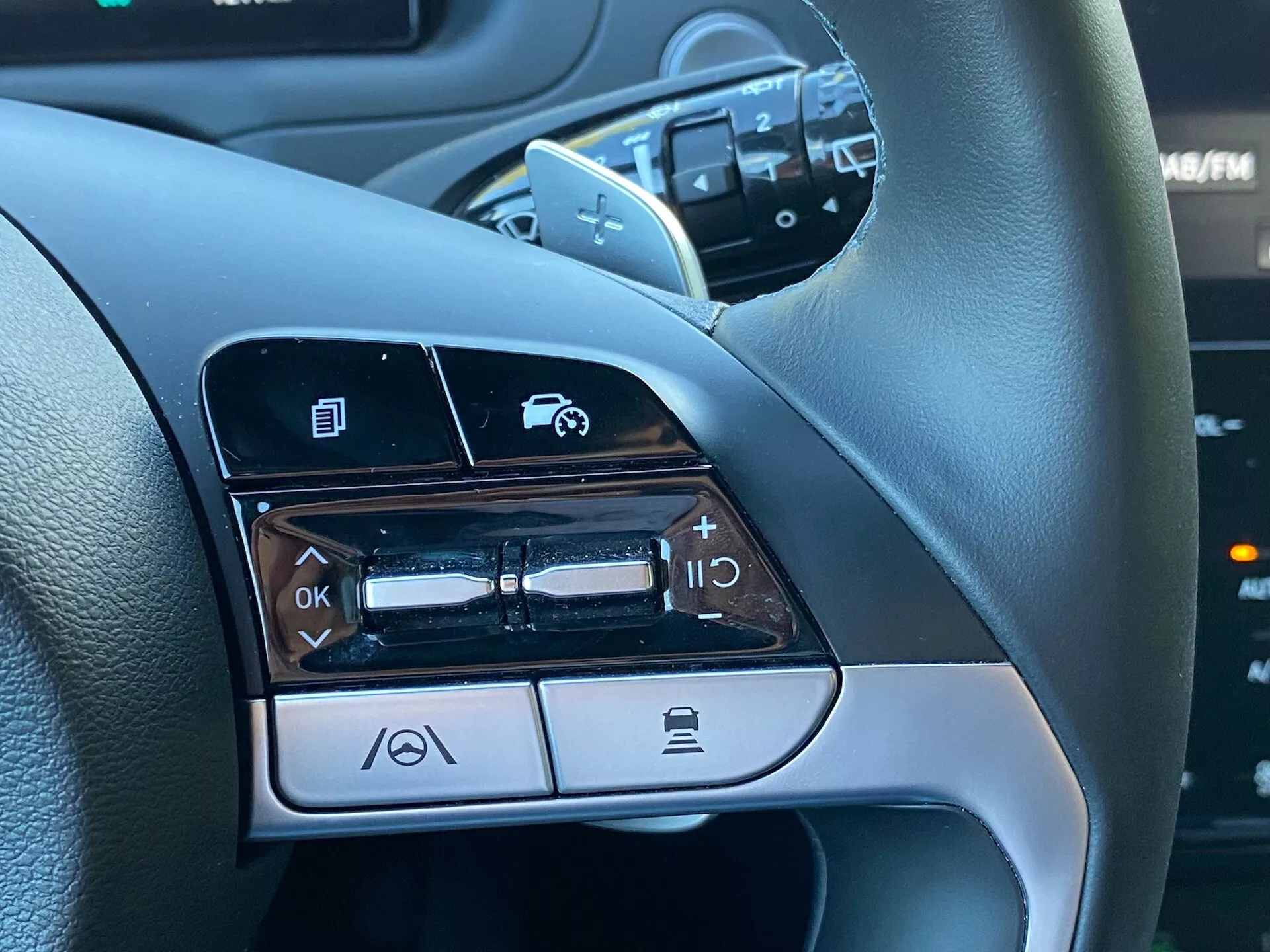 Pogled od blizu na krmilne gumbe na levi strani volana Hyundai Tucson 1.6 T-GDI Pure 2020, ki prikazuje funkcije, kot so navigacija po menijih, nadzor glasnosti, aktivacija glasovnih ukazov in funkcionalnost Bluetooth za telefonske klice.