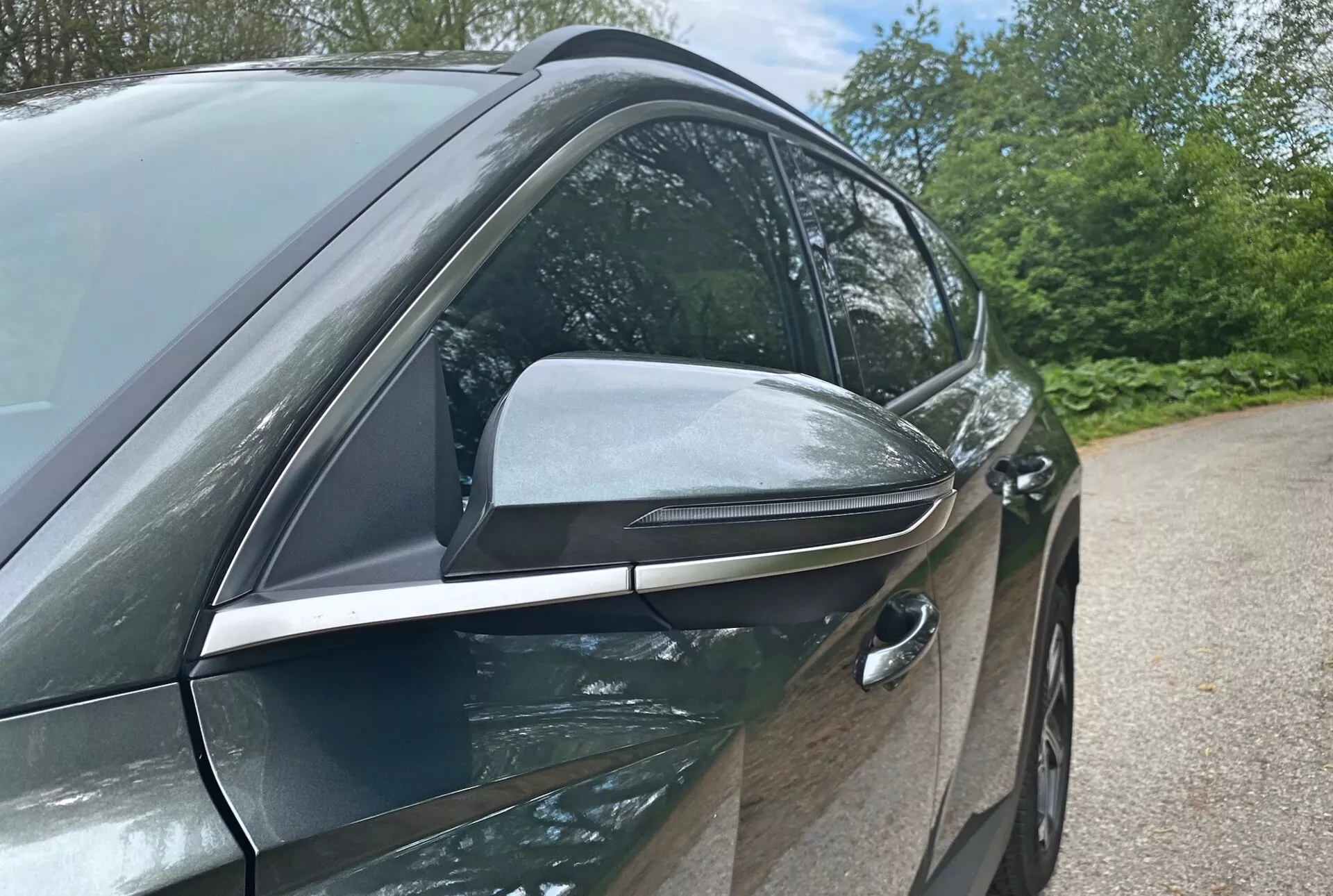 Bližnji stranski pogled na Hyundai Tucson 2020 1.6 T-GDI Pure, ki poudarja njegov moderen dizajn in tehnološke značilnosti s poudarkom na voznikovi strani. Fotografija iz nizkega kota poudarja elegantno, aerodinamično oblikovano stransko ogledalo z vgrajenimi smerniki in črnimi poudarki, kar nakazuje napredne funkcije. Vgrajen sprednji ročaj vrat voznika nakazuje sistem brezklúčnega vstopa, kar prispeva k aerodinamični učinkovitosti vozila. Sijajna, karoserijsko obarvana vrata in kromirani okenski okvirji nakazujejo premium paket opreme. Elementi v ozadju, kot so drevesa in nebo, nakazujejo zunanje okolje.