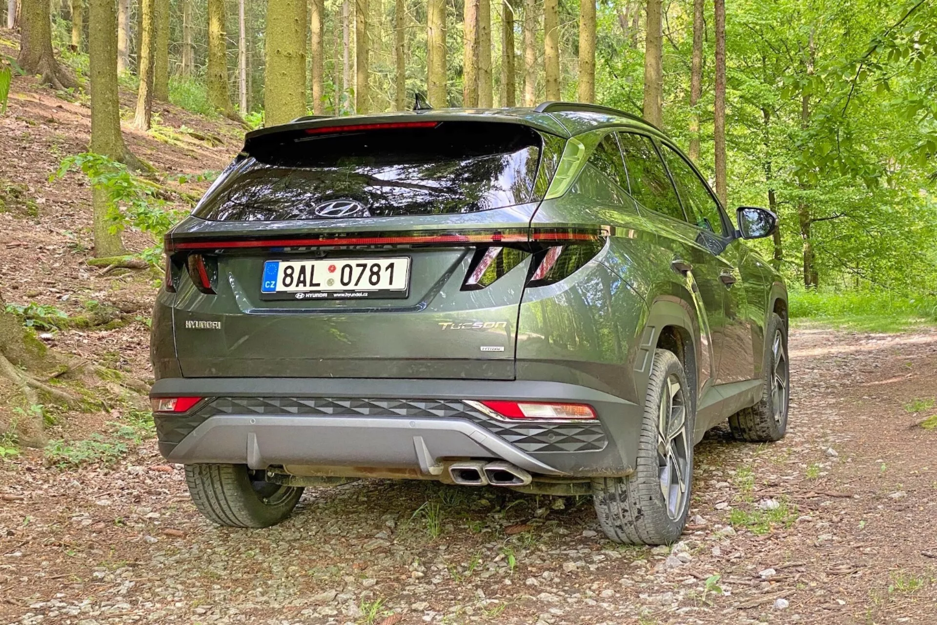 Slika sodobnega SUV-ja Hyundai Tucson 2020 v gozdnem okolju na gramozni poti, posneta iz zadnjega trikvartnega kota. Fotografija poudarja desno stran in zadnji profil vozila z vidnimi zadnjimi vrati, LED zadnjimi lučmi, dvojnim izpušnim sistemom, zadnjim spojlerjem in litimi platišči.