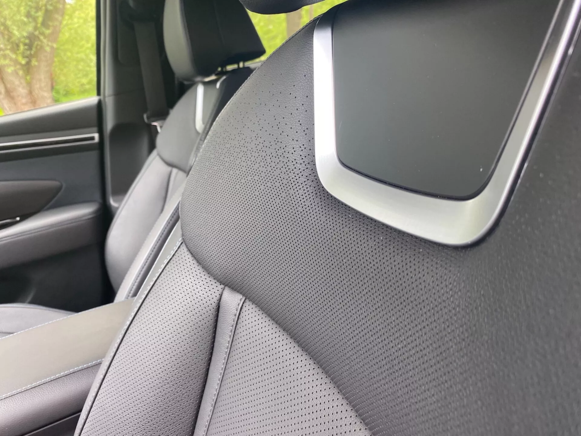 Povečava notranjosti Hyundai Tucson 2020, osredotočena na desni A-stebriček in del desnega sprednjega sedeža. A-stebriček je prekrit z teksturiranim materialom, verjetno tkanino ali sintetičnim usnjem, kar nakazuje poudarek na estetiki ali zmanjšanju hrupa. Sedež ima dvobarvno zasnovo in je perforiran za prezračevanje. Opazna značilnost na stičišču A-stebrička in stropa bi lahko bila del avdio sistema, sestavni del zračne blazine ali senzor za pomoč vozniku, kar poudarja napredne varnostne ali tehnološke funkcije.