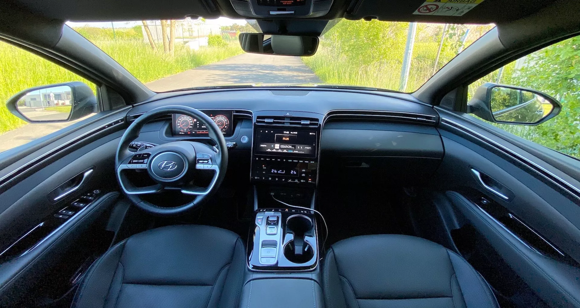 Notranji pogled na Hyundai Tucson 2020 z zadnjega sedeža prikazuje sprednjo armaturno ploščo, volan z vgrajenimi krmilnimi gumbi, digitalni instrumentni panel, velik zaslon infotainment sistema na dotik, gumbe za upravljanje klimatske naprave, prestavno ročico in usnjene sprednje sedeže v naravni dnevni svetlobi.