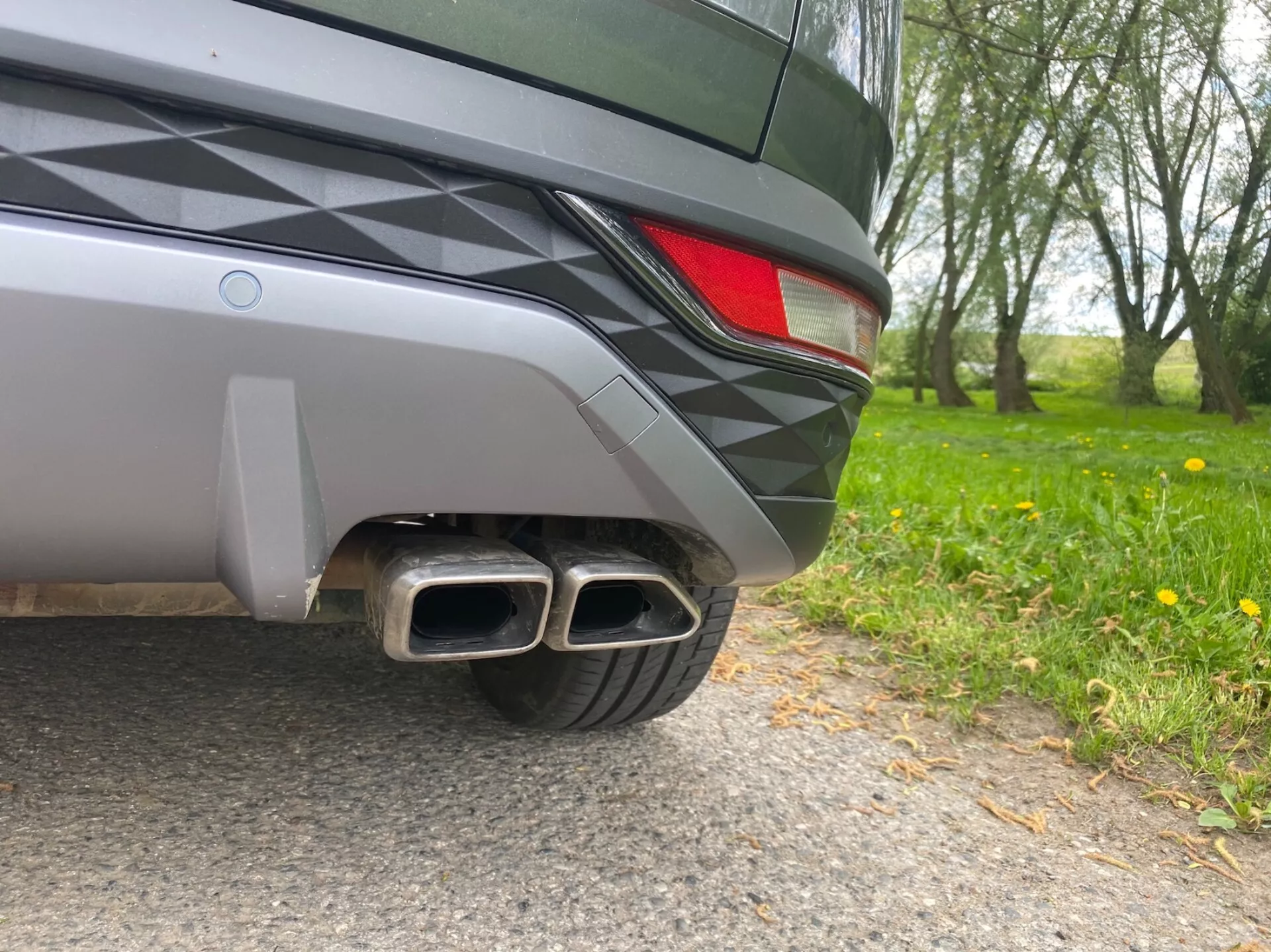 Povečava slike zadnjega dela vozila Hyundai Tucson 2020 z nivoja tal. Vidni detajli vključujejo dvobarvni zadnji odbijač, delno LED zadnjo luč in dvojne kovinske izpušne cevi s pravokotnimi zaključki, kar nakazuje na športno opremo. Elegantna zasnova vozila in kovinska barva odražata naravno okolje.