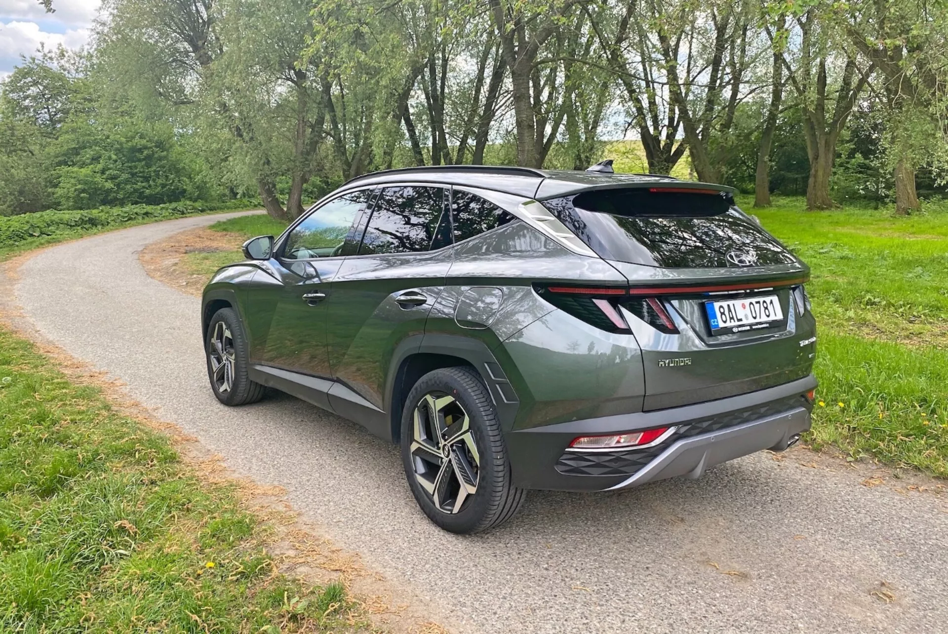 Slika prikazuje sodoben crossover SUV iz zadnjega tričetrtinskega kota, ki poudarja elegantno aerodinamično zasnovo in poševno strešno linijo. Temno zeleno vozilo ima zadnja vrata v slogu liftback, športni spojler in velika lita platišča z večkrakim dizajnom. Vidni elementi vključujejo sodobne zadnje LED luči, dvojne izpušne cevi in ​​zadnji brisalec. SUV je parkiran na gramozni poti, obdani z zelenjem, kar nakazuje park ali podeželsko okolje.
