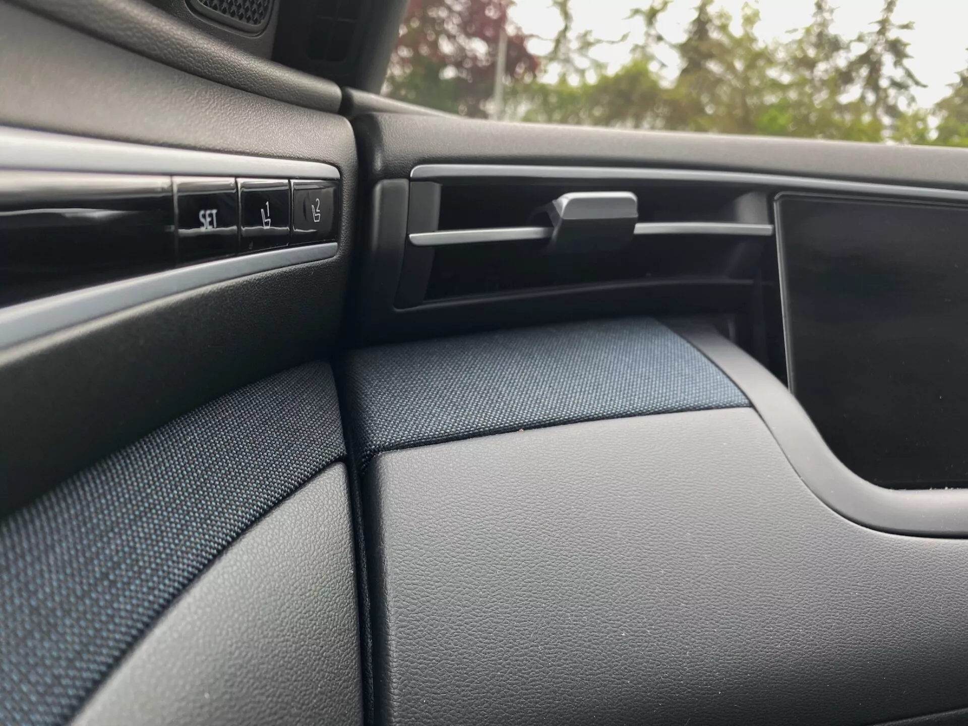 Podroben prikaz notranjosti Hyundai Tucson 2020, ki prikazuje temno, teksturirano stran armaturne plošče na strani sovoznika in del vratne plošče. Scena poudarja več kontrolnih gumbov, nastavljivo zračno odprtino z vodoravnimi letvicami, zaklenljiv predal za rokavice in ukrivljeno zasnovo vratne plošče, ki se brezhibno zlije z armaturno ploščo.