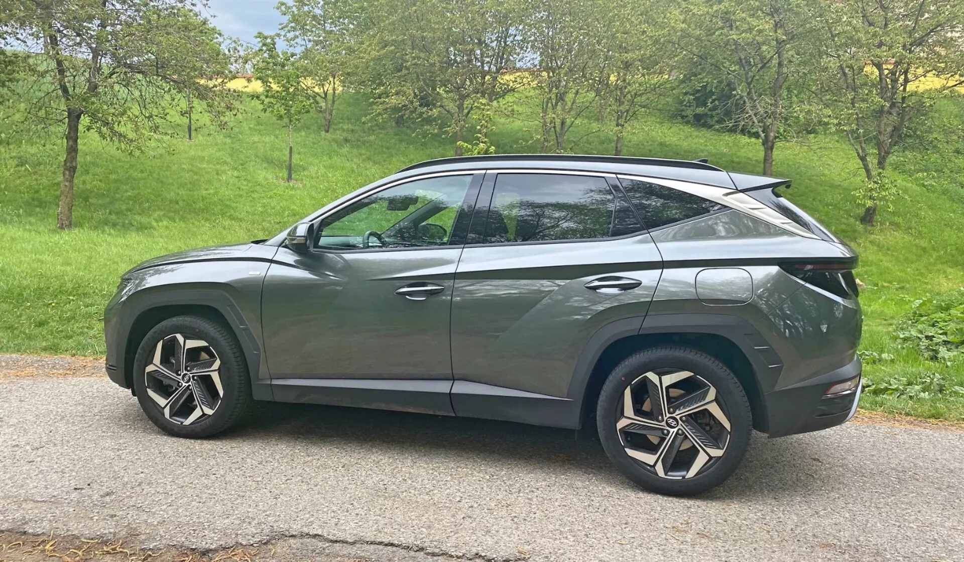 Stranski profil Hyundai Tucson 1.6 T-GDI Pure SUV 2020 v naravni dnevni svetlobi. Vozilo je postavljeno na travnati površini z drevesi v ozadju, kar nakazuje podeželsko ali primestno okolje. Sodobne oblikovalske značilnosti vključujejo LED žaromete, v smerna svetila vgrajena v stranska ogledala, zatemnjena okna, aerodinamična lita platišča in kupejevsko oblikovano poševno streho. Elegantna karoserija z značilnimi linijami in potopljenimi kljukami na vratih nakazujejo napreden brezključni vstopni sistem, medtem ko zadnji spojler kaže na aerodinamične izboljšave. Avtomobil je nameščen v središču okvirja, kar zagotavlja čist, neovirani pogled iz srednje višinske perspektive.