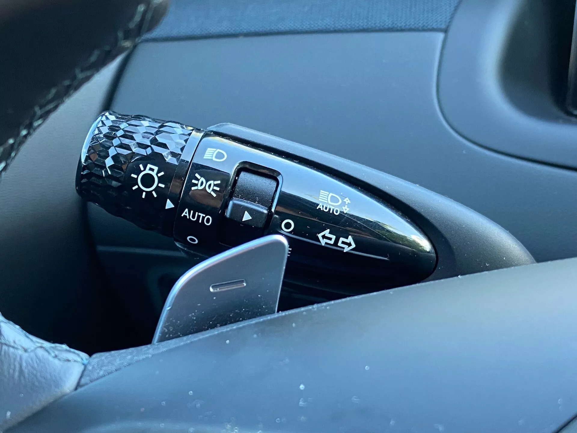 Posnetek od blizu kontrolne ročice, ki se nahaja na volanskem stebru levo od volana v Hyundai Tucson 2020. Ročica vsebuje simbole za nastavitve žarometov, vključno z izklopom, samodejnimi žarometi, parkirnimi lučmi in kratkimi/dolgimi žarometi. Slika prikazuje ročico, del volana in del zamegljenih vrat na voznikovi strani, pri tem pa se osredotoča na podrobnosti in kakovost oblikovanja. Položaj 'AUTO' označuje sistem samodejnih žarometov.