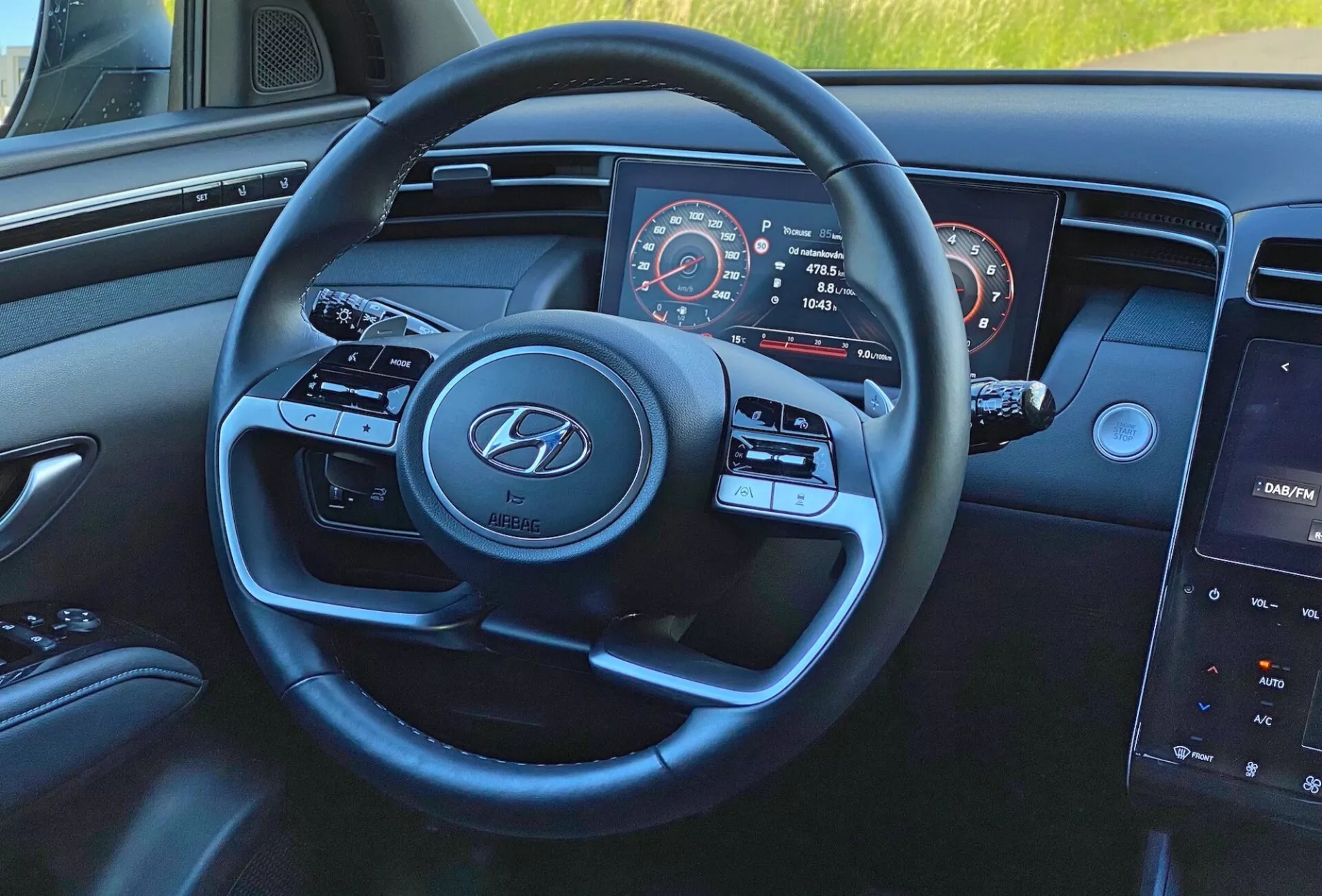 Notranji pogled Hyundai Tucson 1.6 T-GDI 2020 iz voznikove perspektive, osredotočen na sodoben trikraki volan z logotipom Hyundai, gumbe za nadzor medijev in tempomata, delno videno instrumentno ploščo s hitrostnim in tahometrom ter osrednji zaslon na dotik z infotainment sistemom na armaturni plošči.