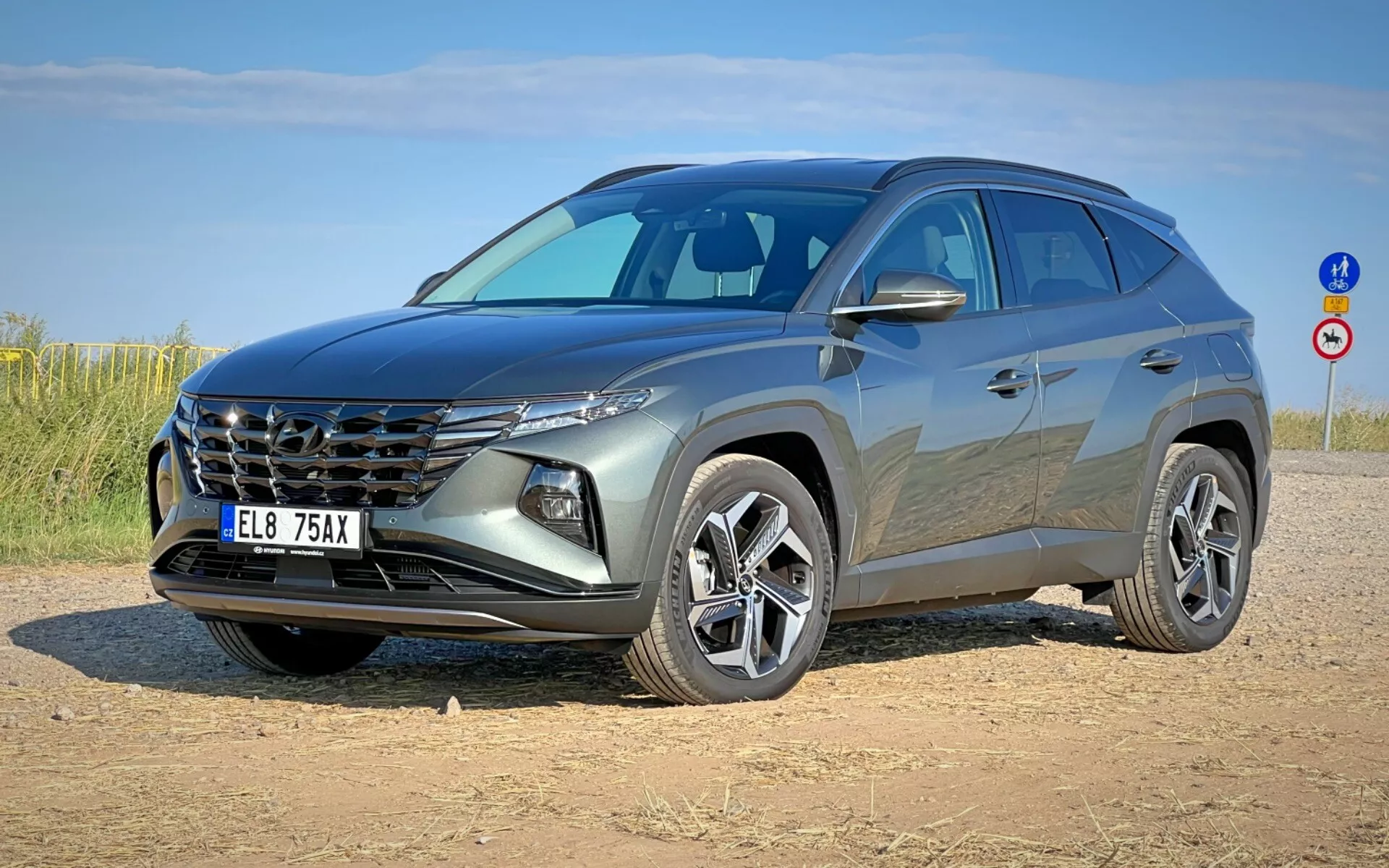 Moderen SUV Hyundai Tucson 2020 prikazan iz treh četrtinskih sprednjih kotov, ki prikazuje njegov značilni žar, elegantne žaromete, sprednji del, kiparsko oblikovano kapuco in lita platišča na makadamskem postajališču pod jasnim nebom
