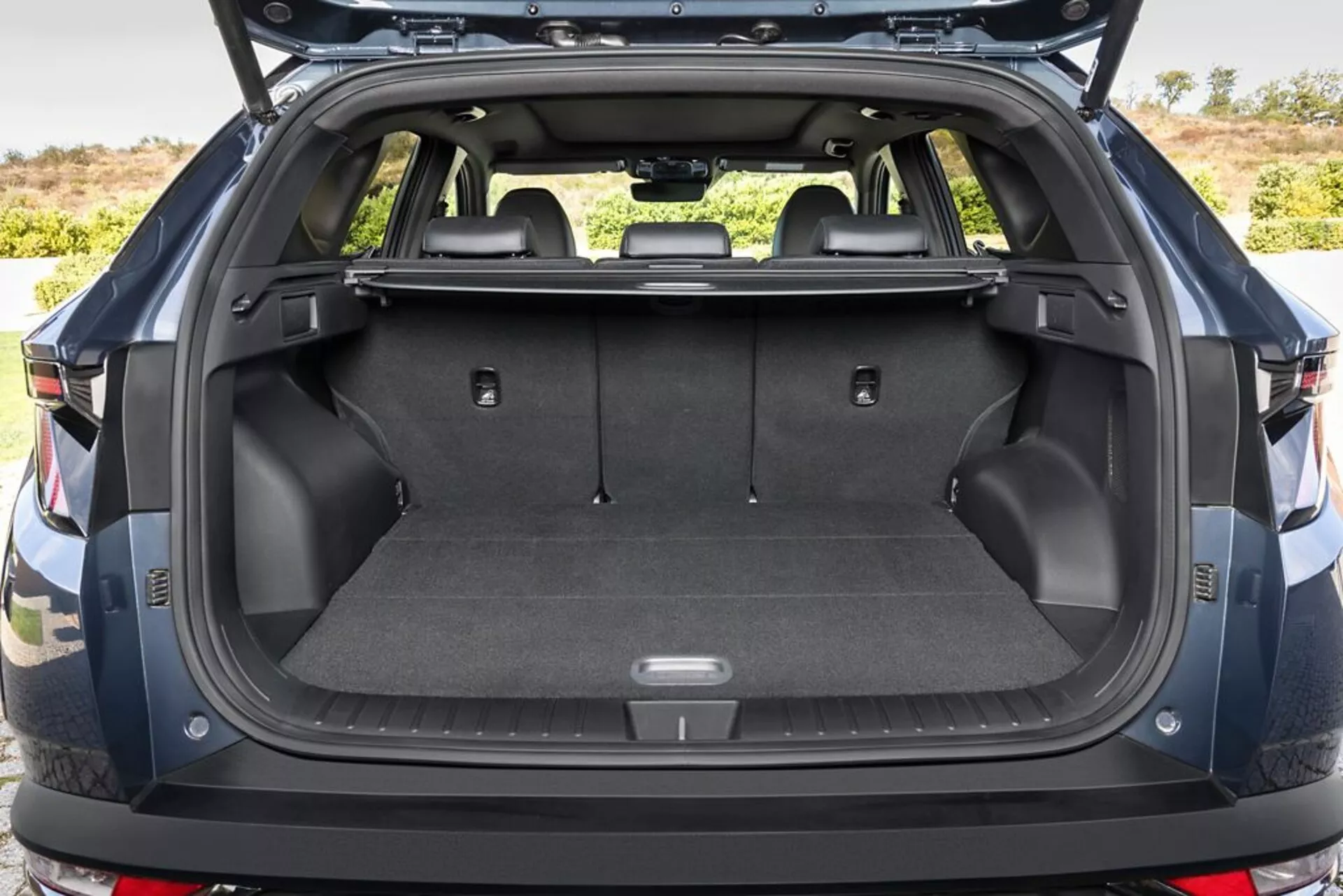 Zadnji tovorni prostor Hyundai Tucsona 2020 z odprtim prtljažnikom, ki prikazuje talno površino, pokrito s preprogo, točke za pritrditev, shranjevalne predele, 12V vtičnico, zložljiva zadnja sedeža, retraktilno zaščito za tovor, vzdržljive plastične stranske stene in delni pogled na zadnji odbijač, na prostem.
