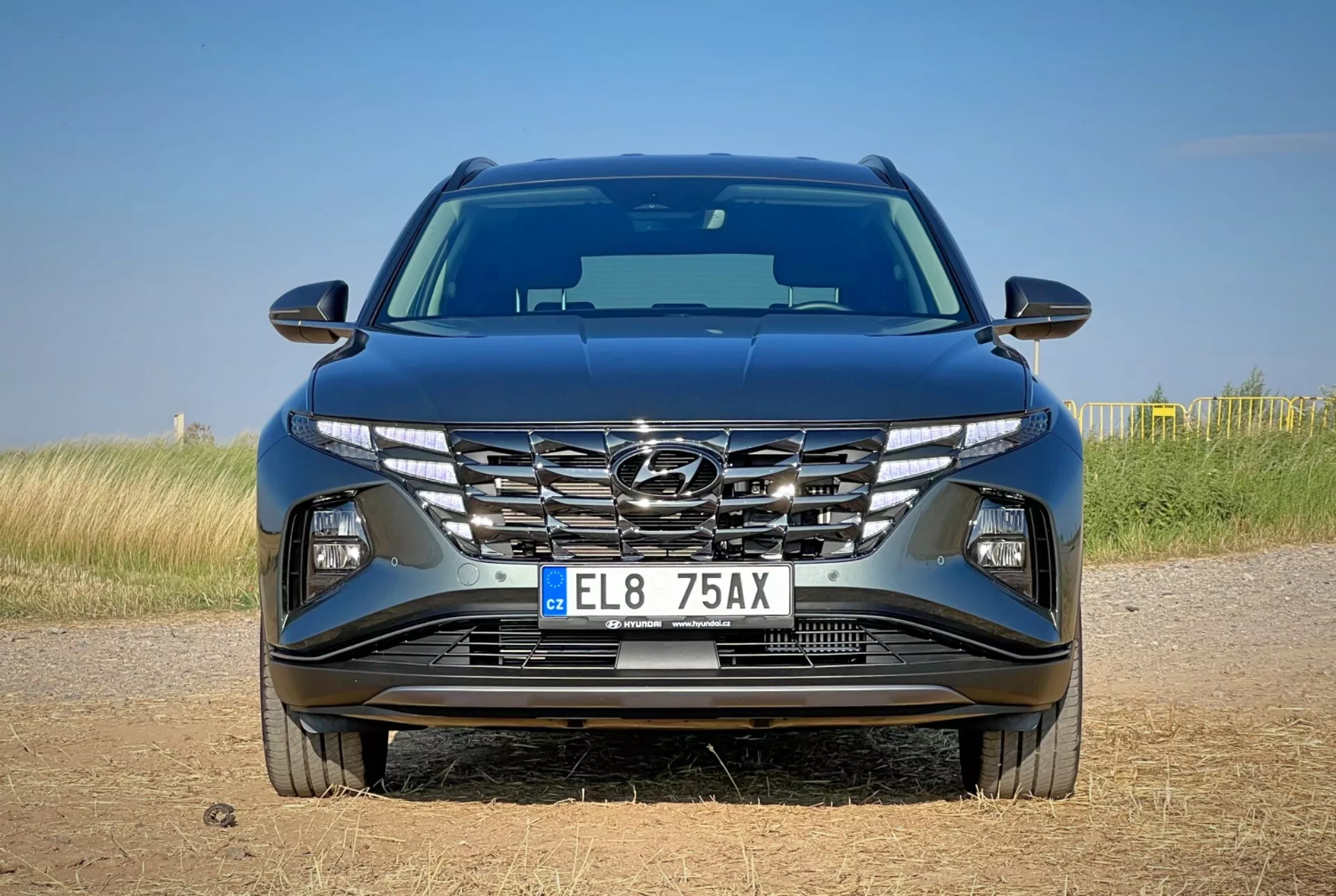 Sprednji pogled na Hyundai Tucson 2020, ki prikazuje aerodinamično obliko pokrova, kaskadno rešetko, ostre LED žaromete in podrobno odbijač z možnimi tehnološkimi funkcijami, postavljen na ozadju naravne pokrajine