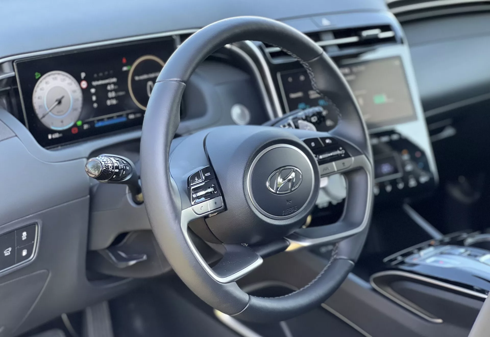 Notranji pogled na Hyundai Tucson 2020, ki se osredotoča na volan in značilnosti armaturne plošče iz perspektive voznika, poudarjajoč trikrako zasnovo z nadzornimi gumbi, digitalni instrumentni sklop in delni pogled na sredinsko konzolo in nadzor klime.