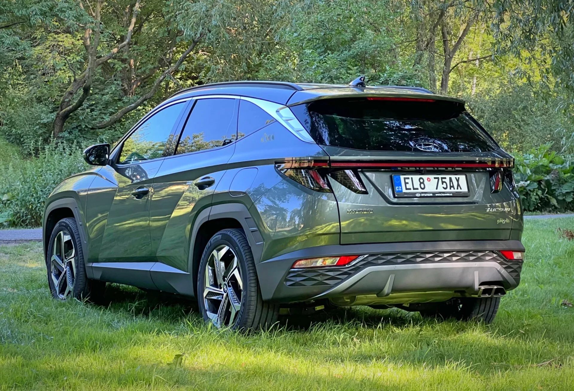 Zadnji tričetrtinski pogled na Hyundai Tucson 2020, ki poudarja LED zadnje luči, parkirne senzorje, dvojne izpušne cevi, športni zadnji spojler in strešne nosilce, parkiran zunaj na travi, prikazuje aerodinamično obliko in napredne funkcije moderne križanke SUV za pustolovščine na prostem in praktično uporabo.