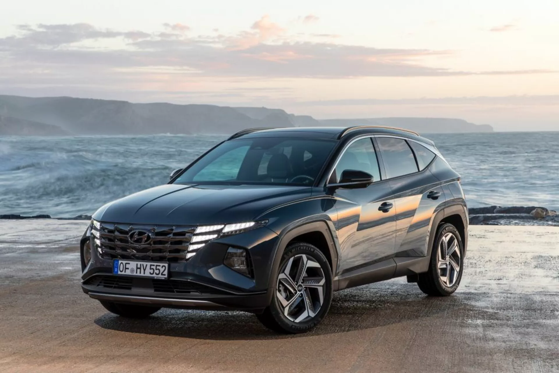 Sodoben SUV Hyundai Tucson 2020 parkiran na obali z ozadjem oceana in obzorja, prikazuje njegov pogled sprednje tri-četrt strani, ki poudarja dizajn vozila, izstopajočo rešetko z logotipom, kotne LED žaromete in športna lita kolesa