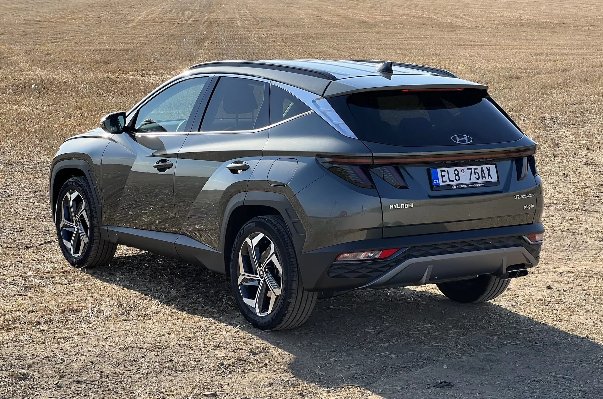 Sodoben SUV Hyundai Tucson 2020 v poševnem pogledu, ki prikazuje zadnje in stranske profile z oznako hibridne tehnologije na suhem travnatem polju, poudarja LED zadnje luči, kontrastni vložek odbijača in športna večkraka kolesa
