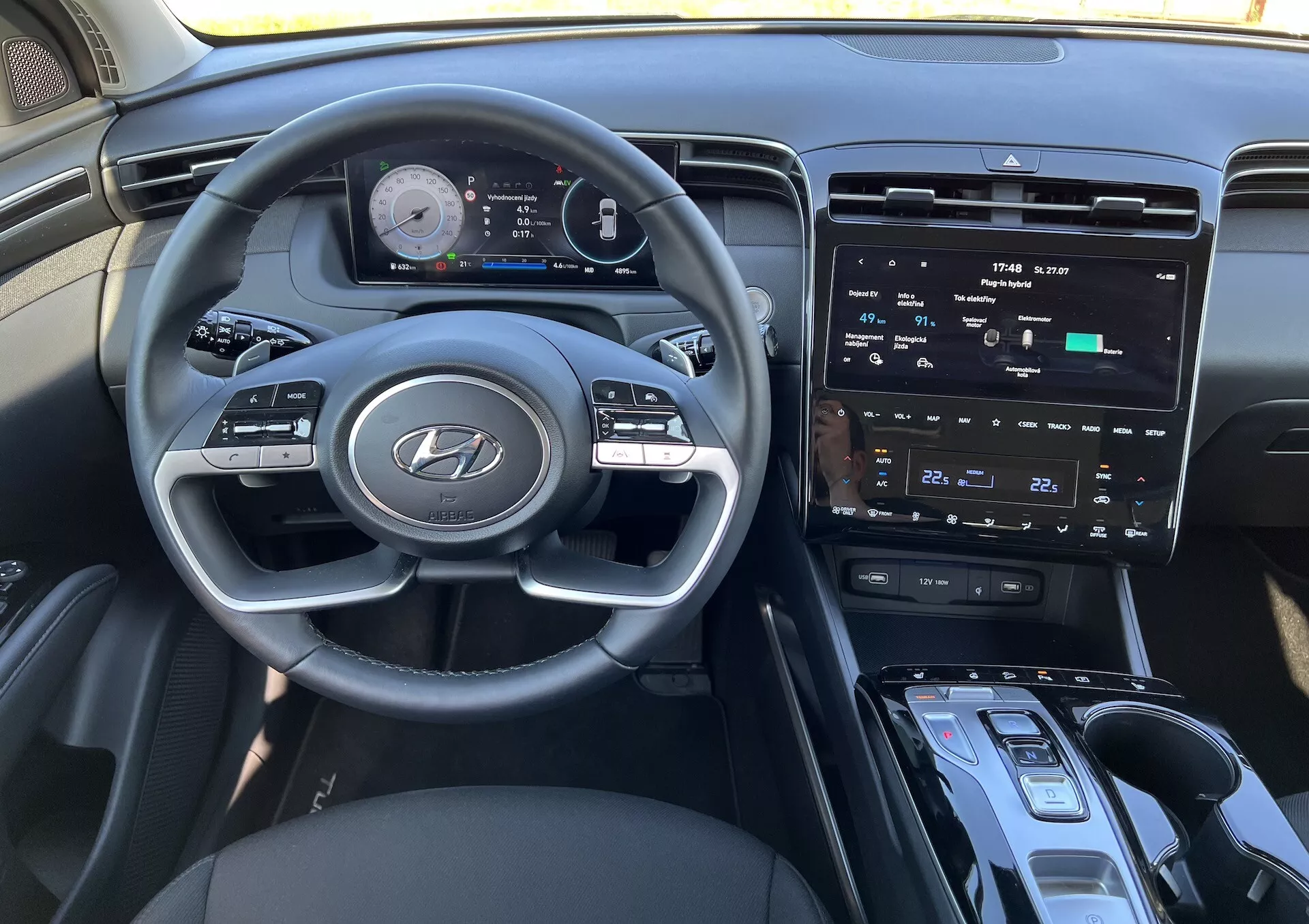 Notranjost Hyundai Tucson 2020 prikazuje volan z logotipom Hyundai, napredno digitalno instrumentno ploščo, centralni zaslon na dotik za informacije in zabavo, kontrole klime, gumb za zagon/zaustavitev, prestavno ročico in možno območje za brezžično polnjenje iz perspektive voznika