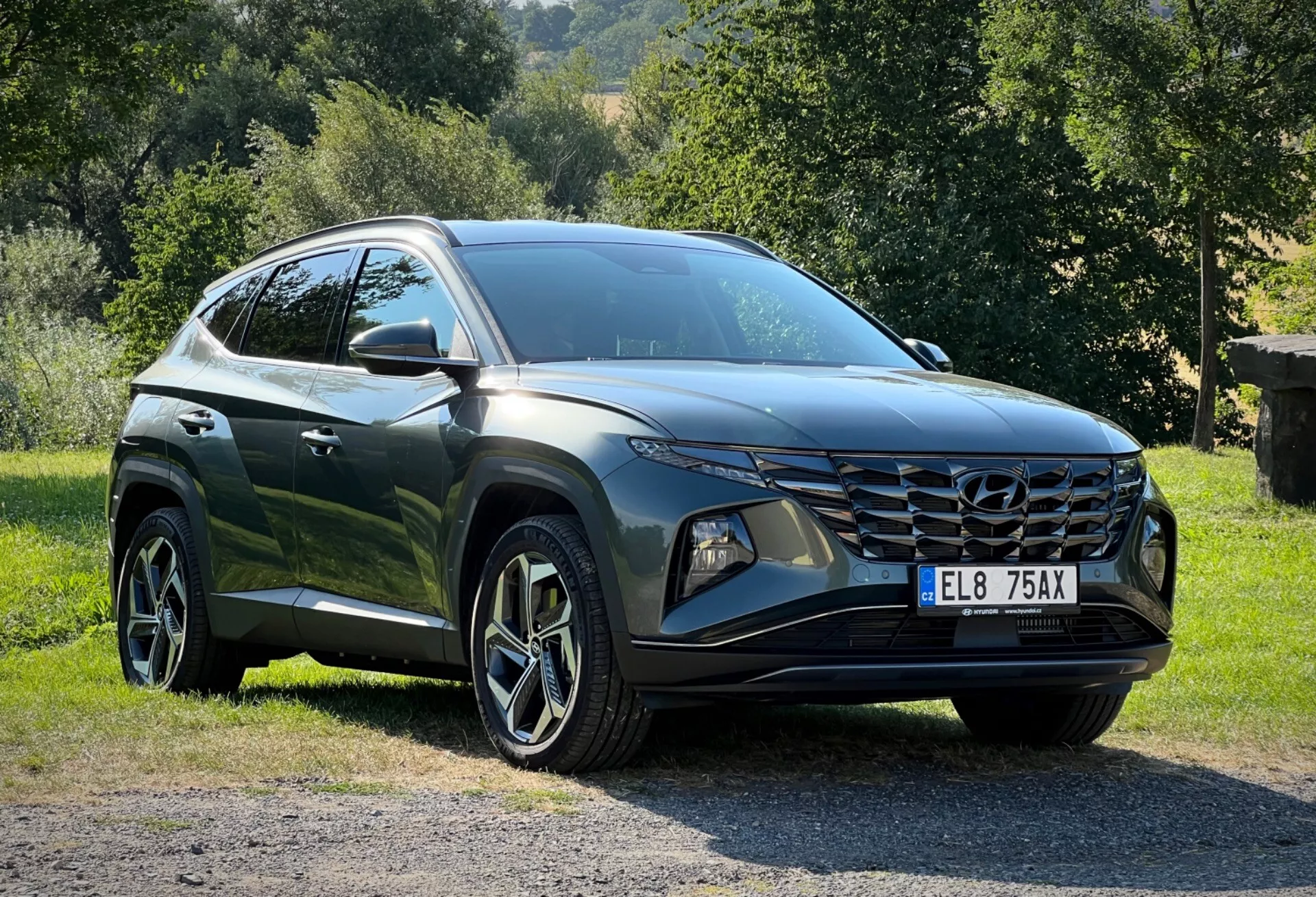 Hyundai Tucson 2020 parkiran na travi z podeželsko kuliso, prikazuje perspektivo s treh četrtin spredaj za podroben pogled na njene elegantne LED žaromete, kotno sprednjo masko in sijočo temno obdelavo