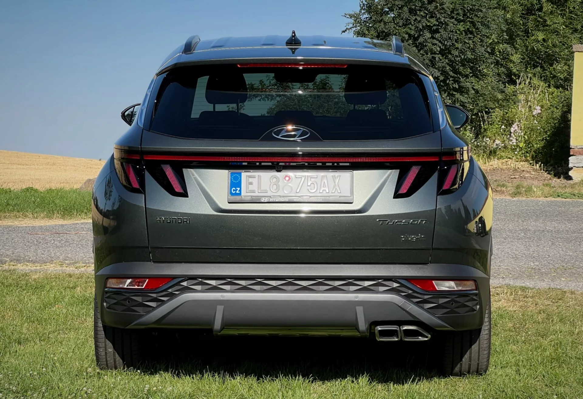 Zadnji pogled na Hyundai Tucson 2020 prikazuje vodoravne zadnje luči, centralni značek, ime stopnje opreme, v odbijač integrirane parkirne senzorje in dvojni izpušni sistem, parkiran na travnati površini proti jasnemu nebu