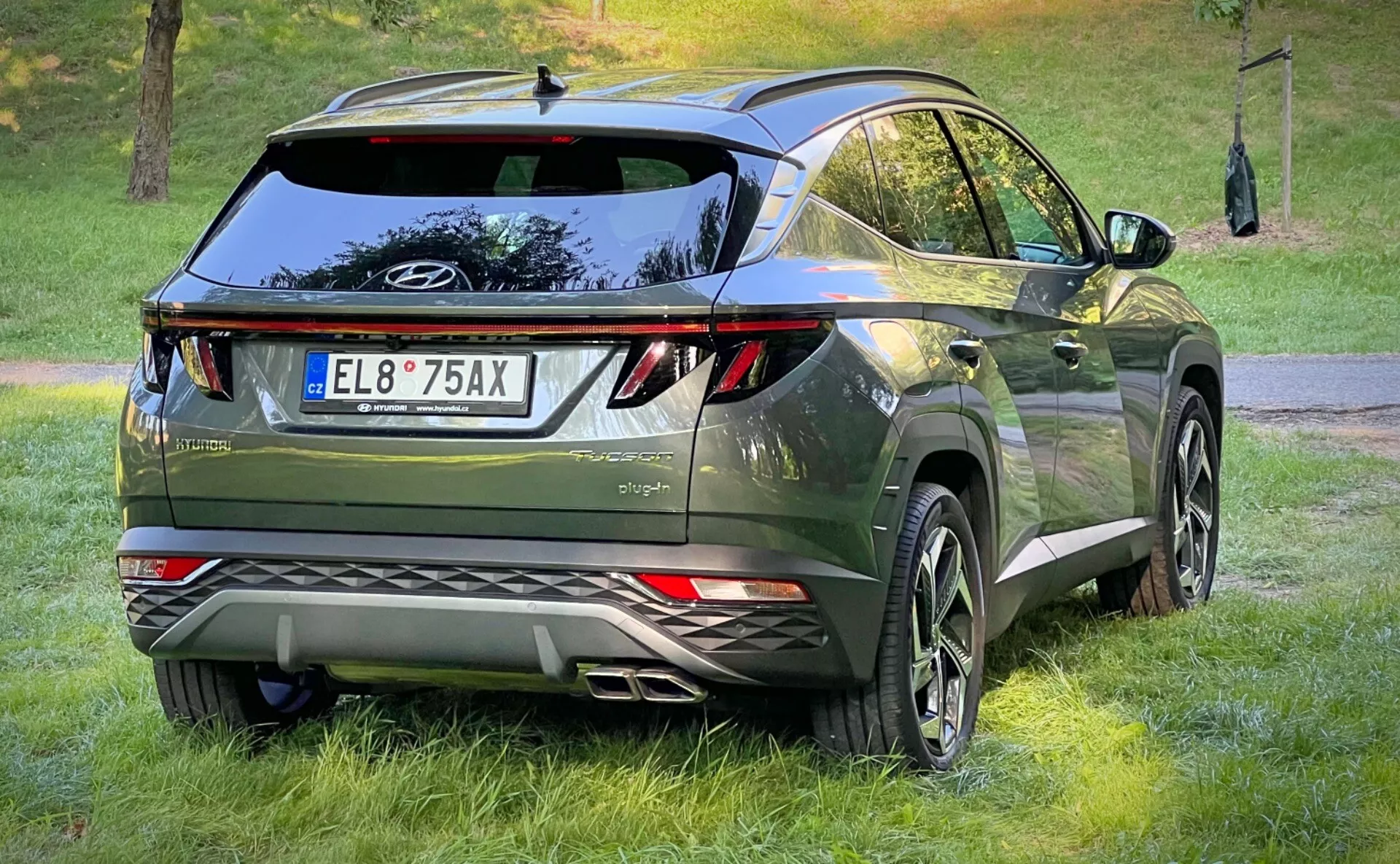 Tričetrtinski zadnji pogled na Hyundai Tucson 2020, ki prikazuje njegove LED zadnje luči, aerodinamično obliko z zadnjim spojlerjem in anteno v obliki morskega psa, premijsko temno zeleno kovinsko dokončanje in opremljen z velikimi aluminijastimi kolesi in senzorji za parkiranje, v urbani crossover življenjski okolici