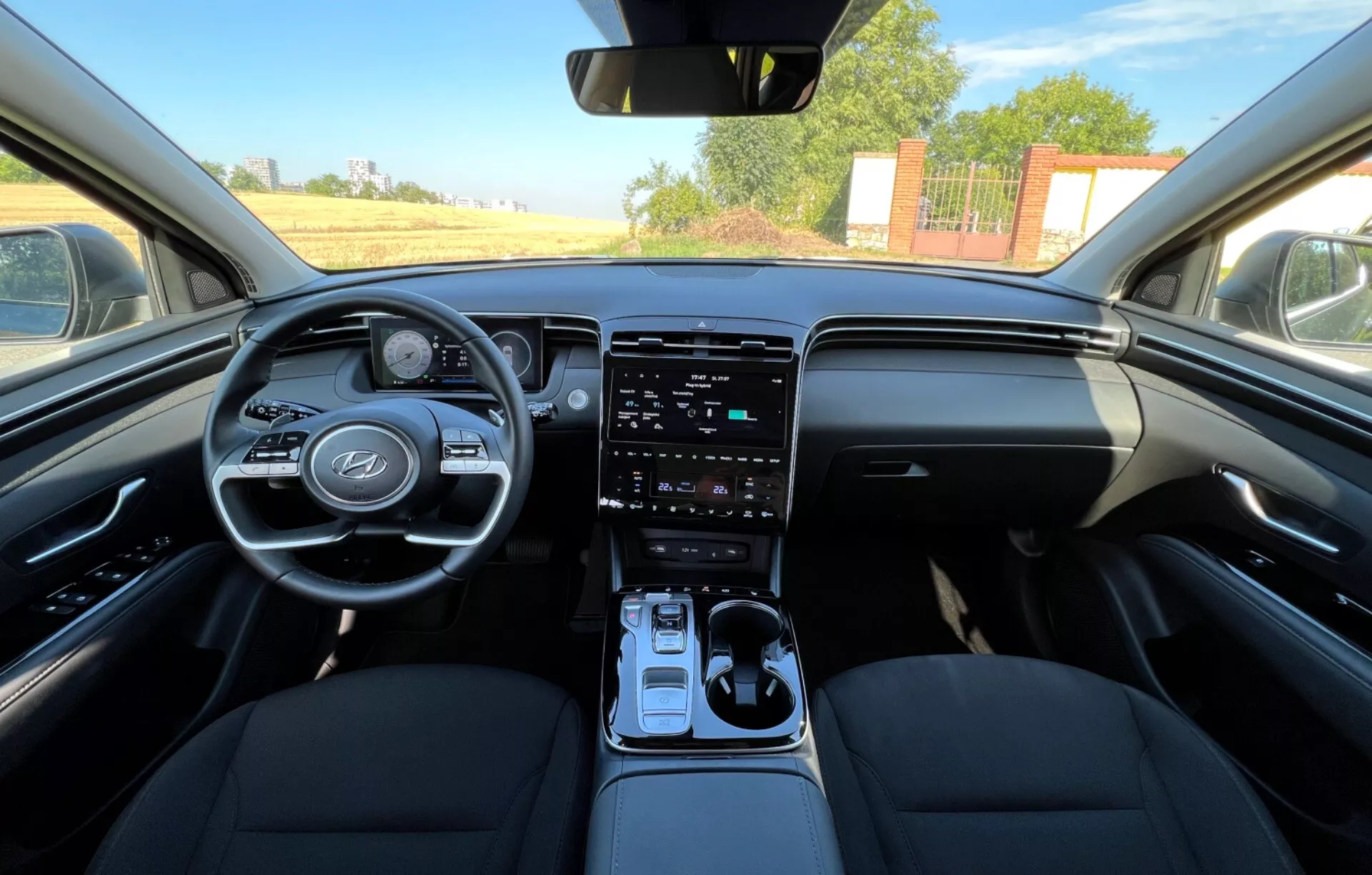 Notranjost Hyundai Tucson 2020 s prikazom sprednjih sedežev, volana z logotipom proizvajalca in kontrolnimi gumbi, digitalnega instrumentnega sklopa, vertikalnega zaslona infotainment sistema na sredinski konzoli, avtomatske prestavne ročice in dvo-tonsko zasnovo armaturne plošče iz perspektive, ki spominja na pogled voznika ali sovoznika