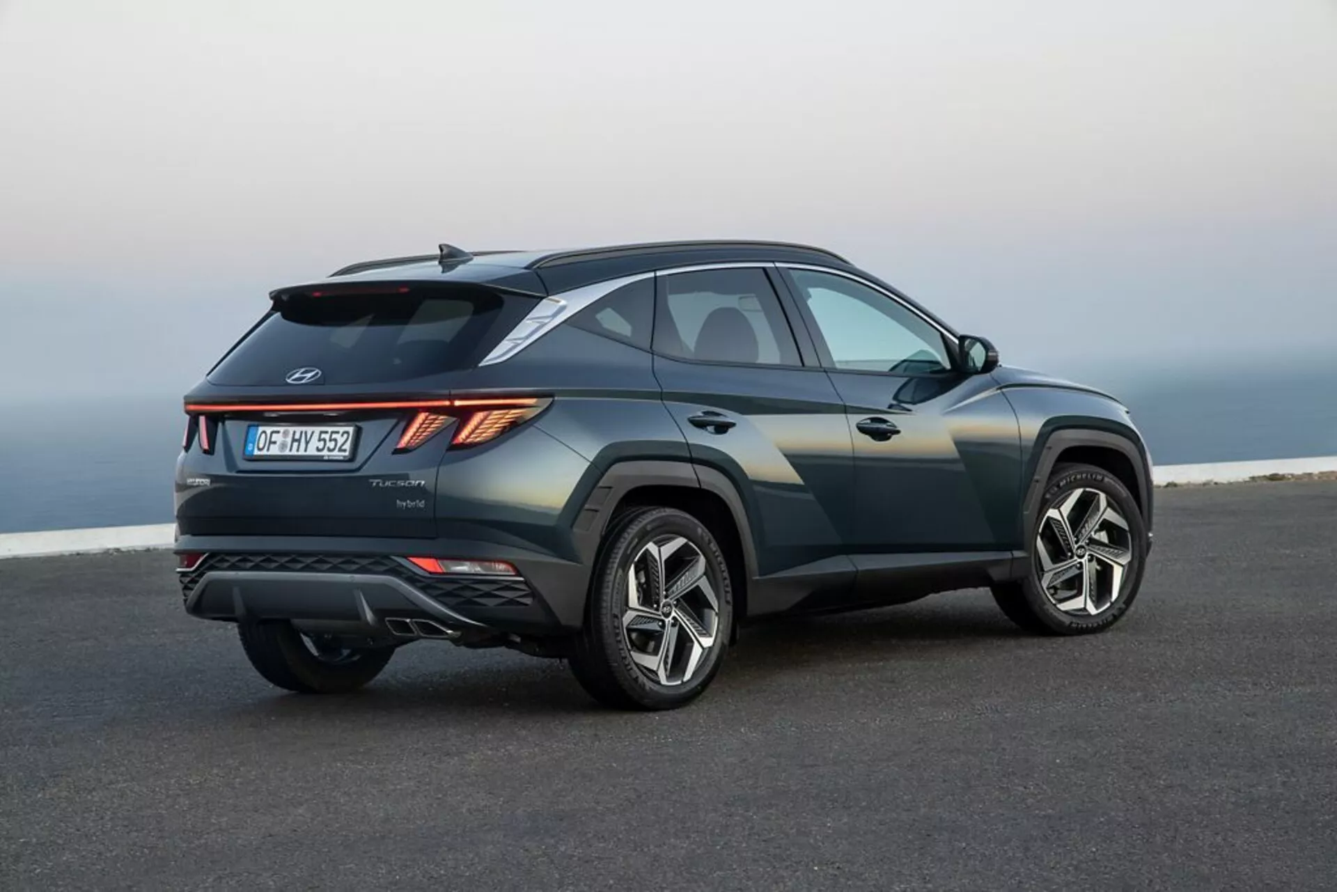 SUV Hyundai Tucson 2020 parkiran na gladkem asfaltu z obzorjem obalne ali visoke pokrajine v ozadju, ki prikazuje njegov eleganten, športen dizajn, naraščajočo linijo pasu, spuščajočo se linijo strehe, ostre robnike karoserije, kotne LED zadnje luči, zadnji spojler, integrirani difuzor, dvojne izpušne cevi in velika lita kolesa