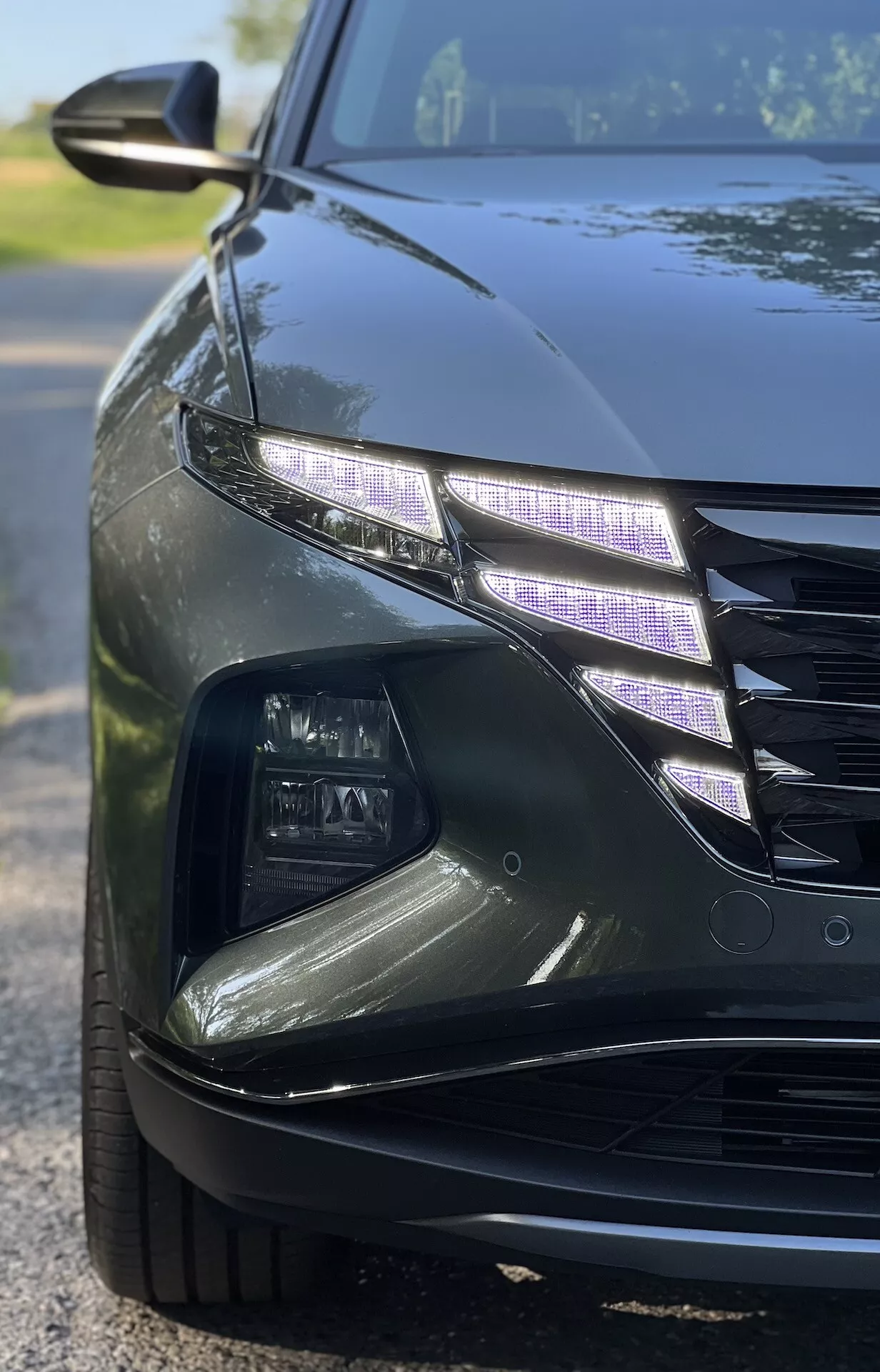 Iz bližnje kotne perspektive Hyundai Tucson 2020 prikazuje zapleten dizajn LED žarometov in del rešetke s sijajnim zaključkom v šibki svetlobi, kar ilustrira lastnosti luksuznih vozil in sodobno tehnologijo avtomobilske razsvetljave.