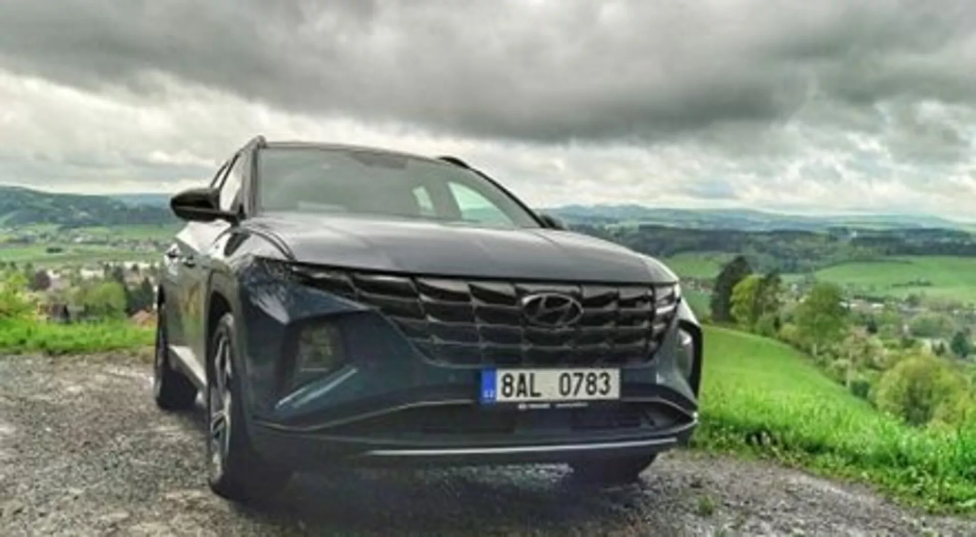 SUV Hyundai Tucson 2020 parkiran na gramozni cesti, poudarja svojo drzno rešetko, elegantne LED žaromete in kovinsko barvo v dinamičnem pogledu iz nizkega sprednjega kota, z bujno hribovito pokrajino pod oblačnim nebom v ozadju