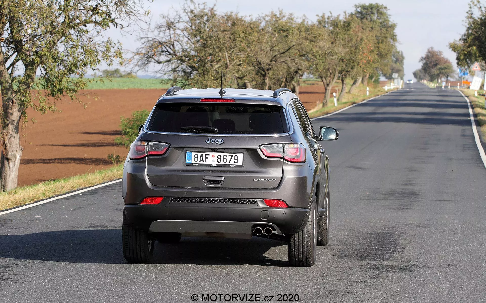 Pogled od zadaj na Jeep Compass 2017, ki se oddaljuje po dvopasovni cesti, prikazuje moderne oblikovalske elemente, kot so antena v obliki plavuti morskega psa, zadnji brisalec, LED zadnje luči, vgrajeni parkirni senzorji, dvojne izpušne cevi in evropsko registersko tablico z jasno vidnim logotipom Jeep. Vozilo se zdi v gibanju, kar zajema resnično delovanje z možnimi tehnologijami za pomoč vozniku.