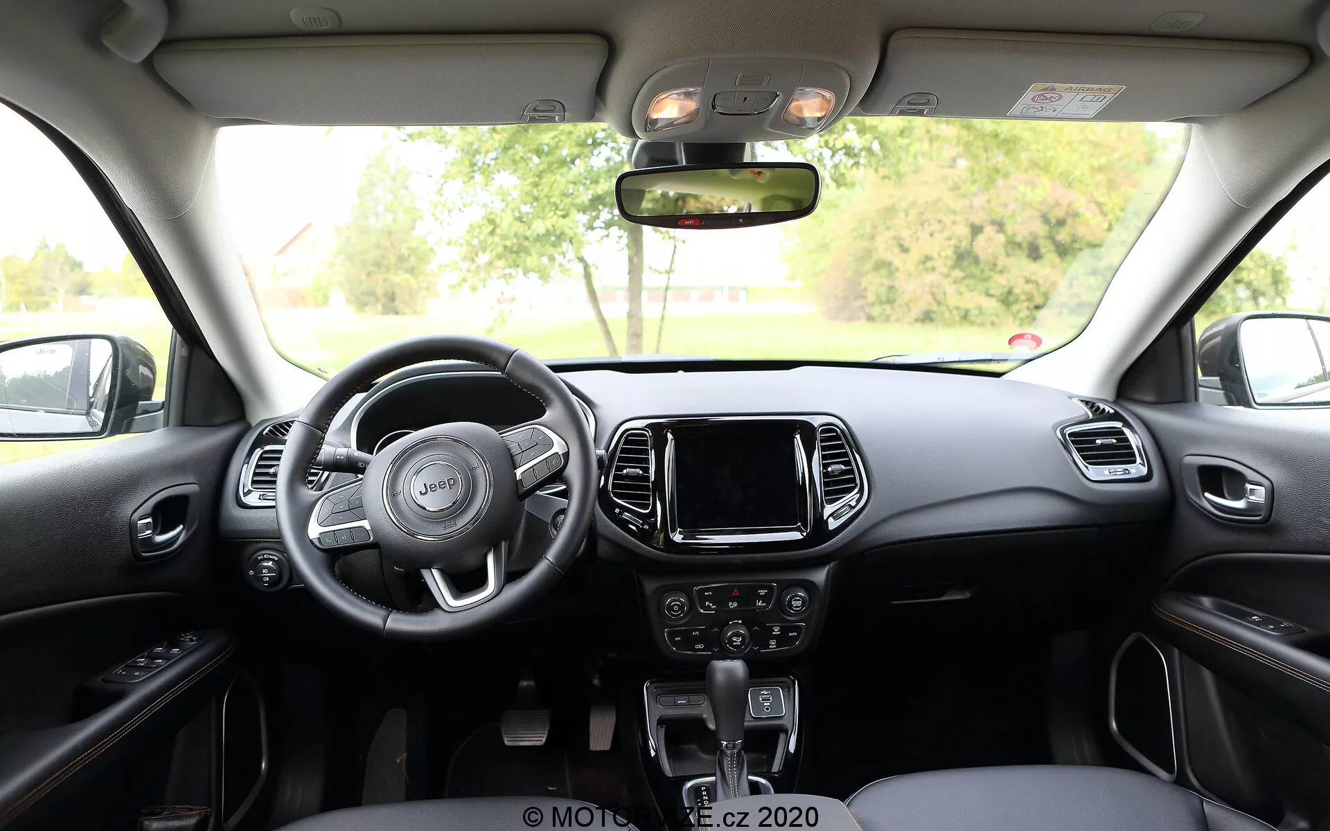 Pogled v notranjost Jeep Compass 2017 iz zadnjega sedeža, prikažuje trikraki volan z integriranimi upravljalnimi gumbi, osrednjo konzolo z zaslonom na dotik za infozabavni sistem, ročne kontrole za avdio in klimo pod njim ter ročico avtomatskega menjalnika. Armaturna plošča in sprednji sedeži imajo dvobarvno shemo s kovinskimi ali plastičnimi poudarki, nad glavo pa so vidni kontrolni gumbi za strešno svetlobo in strešno okno.