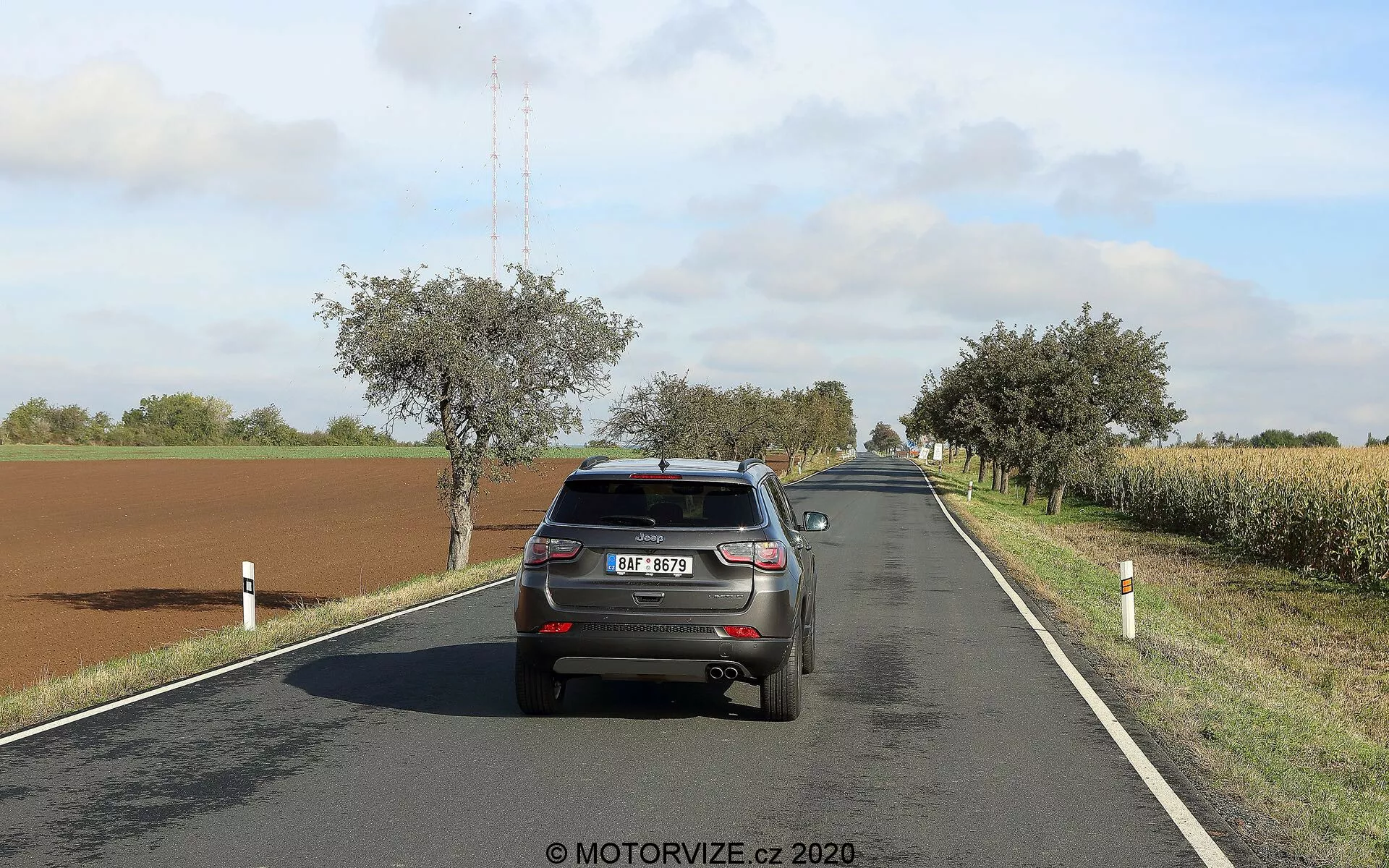 Slika prikazuje zadnji del Jeep Compass 2017, ki vozi po dvopasovnici podeželski cesti na sončen dan z razpršenimi oblaki. Vozilo, ki je nameščeno na desni strani pasu, prikazuje svoja zadnja vrata, odbijač, zadnje luči, zadnje vetrobransko steklo, registrsko tablico, del strehe s anteno v obliki plavuti morskega psa in zgornji del zadnjih pnevmatik. Avto zaseda pomemben del okvirja, kar omogoča jasno prikaz njegovih lastnosti. Cesta se v daljavi nežno zavija v desno, obdana s polji, drevesi in cestnimi drogovi. Zavorevne luči avtomobila niso prižgane, kar nakazuje na normalno vožnjo.
