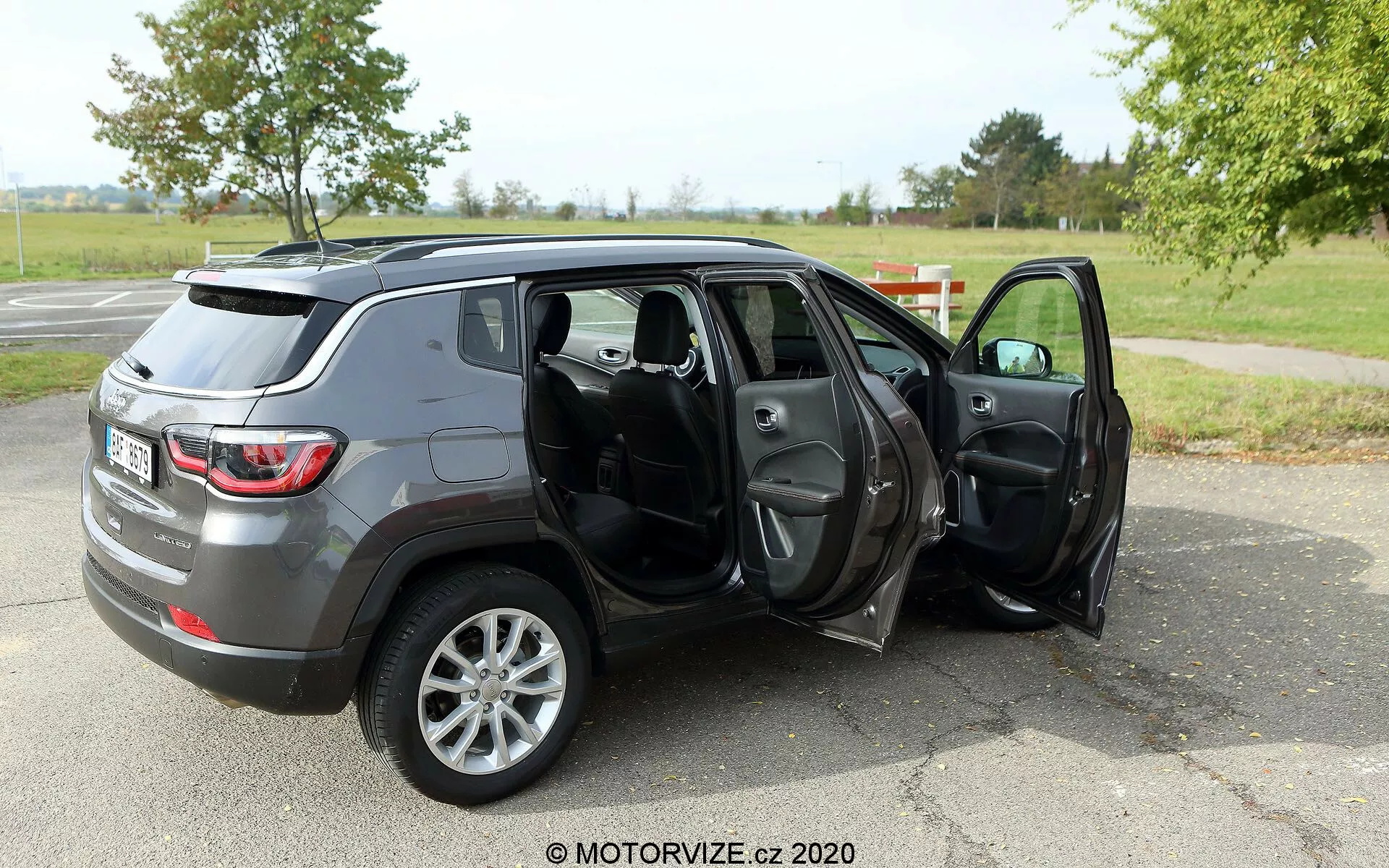 Slika športnega terenca Jeep Compass 2017 srednje velikosti, parkiranega na asfaltni površini v okolju, ki spominja na park s travo in drevesi v ozadju. Fotografija je posneta iz zadnjega trikotnega kota, ki prikazuje profil in zadnji del vozila. Obe potniški vratni sta odprti, kar razkriva notranjost iz temnega blaga ali usnja, vključno s sprednjimi in zadnjimi sedeži, s potencialnimi lastnostmi, kot so električno upravljana okna in ogledala. SUV ima siv kovinski zaključek in je opremljen s petkrakimi aluminijastimi kolesi, kar lahko nakazuje višjo stopnjo opreme ali opcijsko nadgradnjo koles. Vodni žig 'MOTORVIZE.cz 2020' nakazuje, da slika izhaja iz recenzije avtomobila ali avtomobilistične spletne strani.