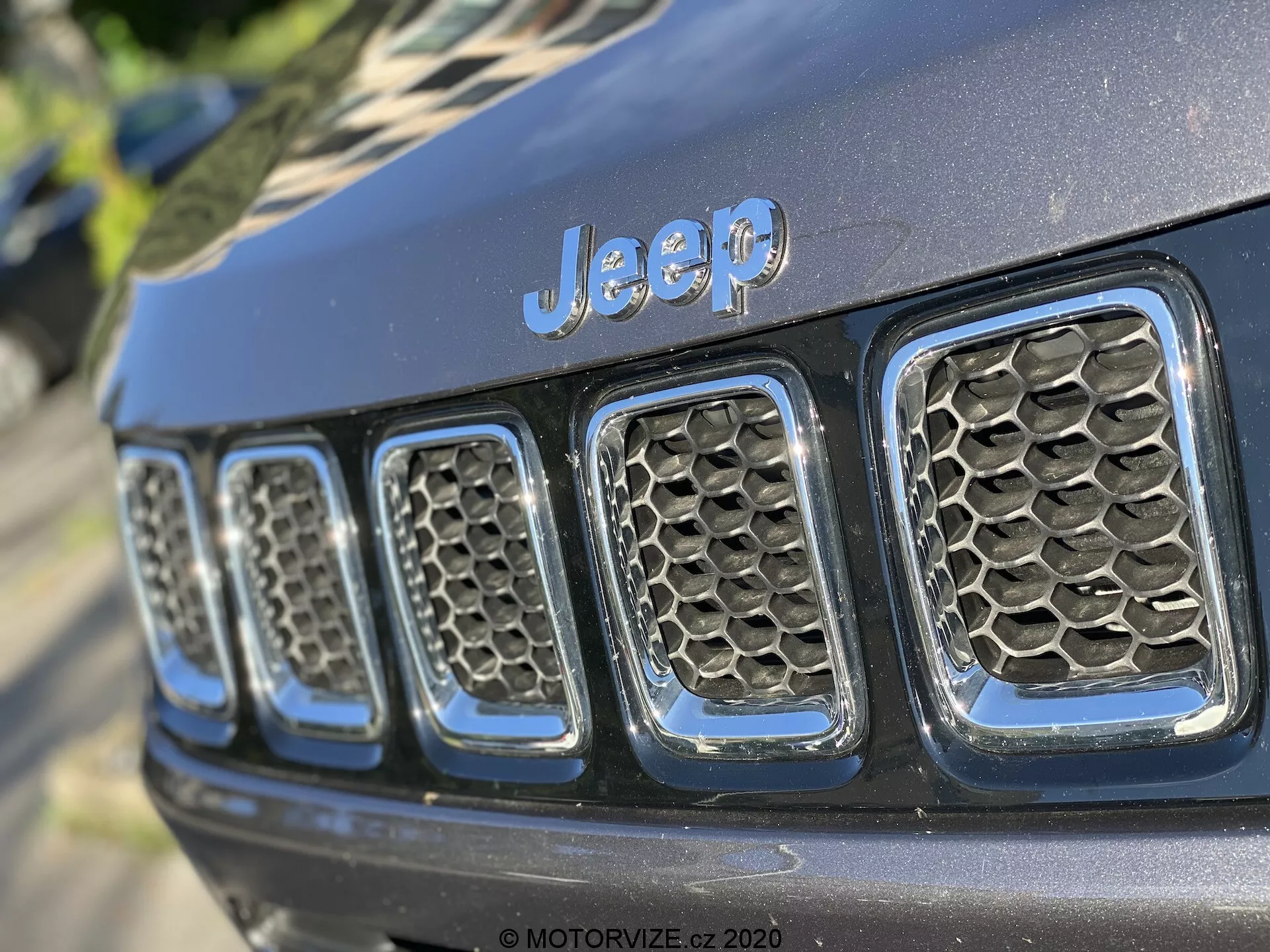 Pogled od blizu na sprednjo rešetko Jeepa, z vidnim prepoznavnim znakom 'Jeep' nad sedmimi navpičnimi režami, kar je značilna oblikovna poteza. Fotografija je posneta pod kotom, osredotoča se na levo stran rešetke in je posneta iz rahlo nizkega kota. Reže rešetke imajo vzorec satovja, kar kaže na izboljšan pretok zraka in zaščito hladilnika in motornih komponent. Sijajna črna barva kaže na razmeroma novo ali dobro vzdrževano vozilo. Plitka globina polja drži znak 'Jeep' in nekaj rež vizualno ostrih, medtem ko ostalo postopoma izostaja, kar poudarja oblikovne in blagovne elemente pod naravno zunanjo svetlobo z mehkimi sencami.