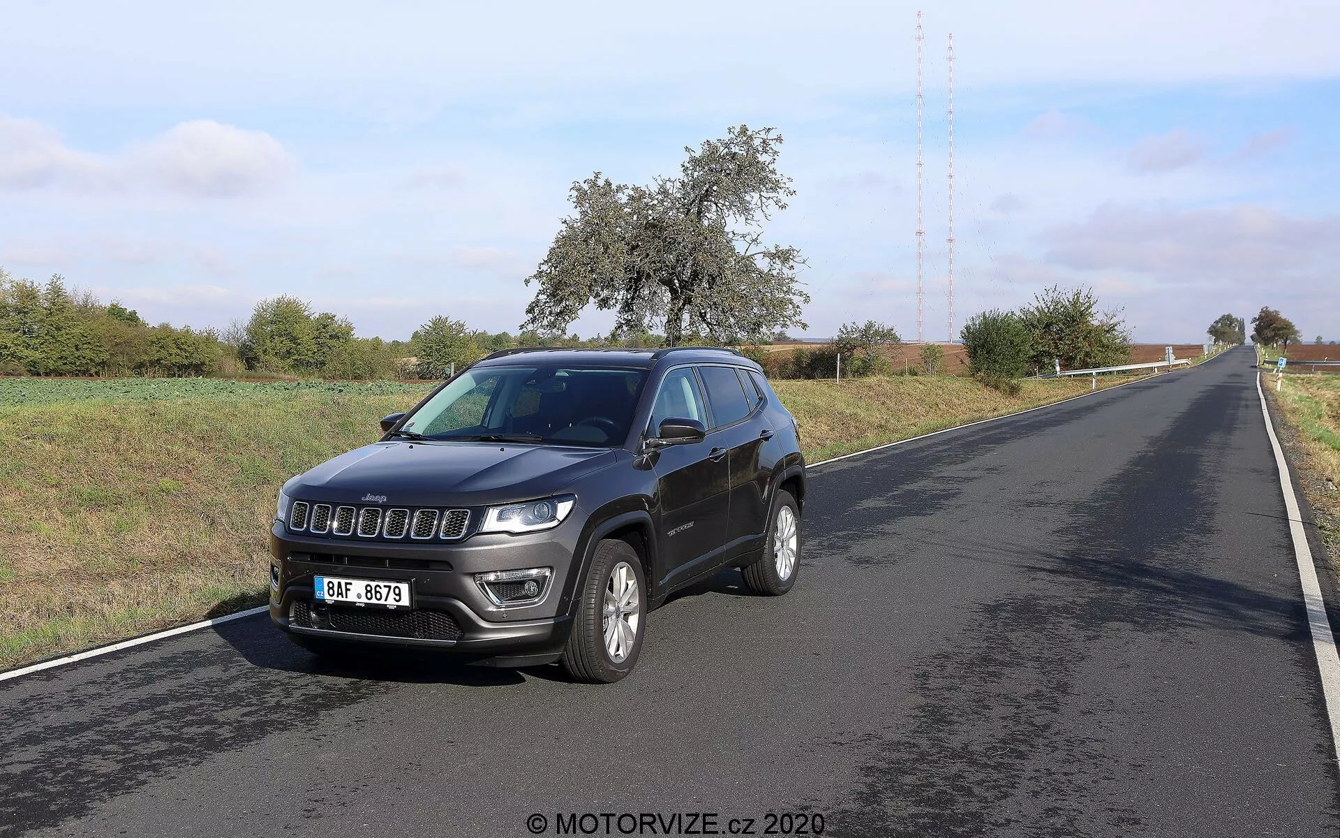 Slika Jeep Compass 2017, fotografirana iz trikotnega sprednjega kota. Vozilo, postavljeno na desni strani dvopasovne ceste, prikazuje svoj sprednji in stranski profil. Ključne značilnosti vključujejo sodobno rešetko, izrazite žaromete z LED dnevnimi lučmi, aluminijasta platišča, integrirane smerne utripalke na stranskih ogledalih, strešno anteno in prednje meglenke. Okolje se zdi podeželsko s čistim nebom in vegetacijo ob cesti.