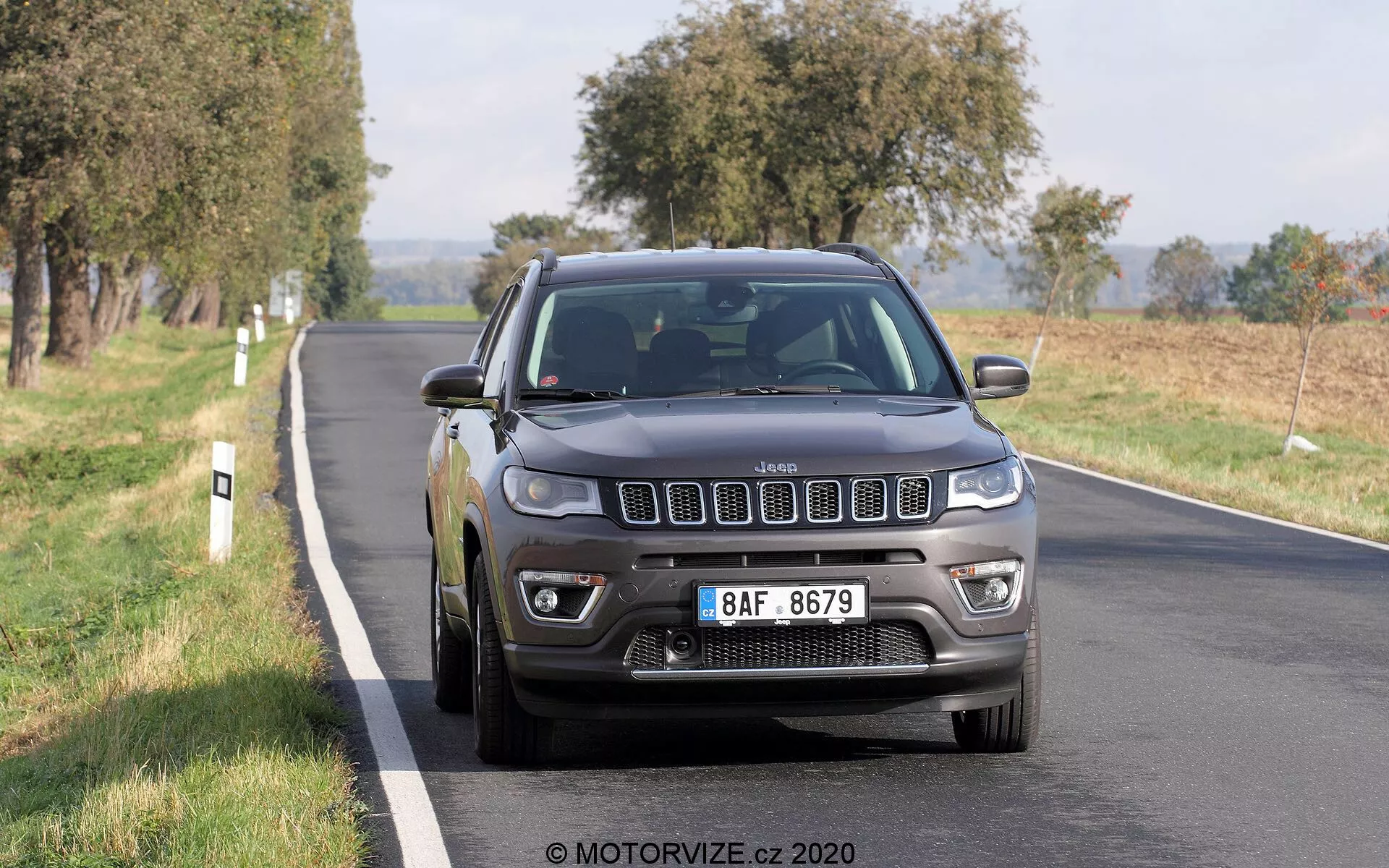 Podoba leta 2017 Jeep Compass, ki se vozi po dvopasovni cesti, fotografirana iz prednjega tridčetrtinskega kota. Ikonična sedem-režna mreža vozila, podrobni žarometi z LED dnevnimi lučmi, meglenke in večkraka aluminijasta platišča so jasno vidni. Temno siva ali črna barva Jeepa mu daje trden in robusten videz, pri čemer naravna dnevna svetloba poudarja njegove značilnosti. Vozilo se zdi v gibanju, s rahlo zameglitvijo koles. Drevesa in odprta polja v ozadju nakazujejo podeželsko ali pol-podeželsko okolje. Avto zaseda srednjo in spodnjo polovico kadra, rahlo desno, pri čemer zajema sprednjo mrežo, žaromete, odbijač, pokrov motorja, voznikov blatnik, kolo in sprednja vrata.